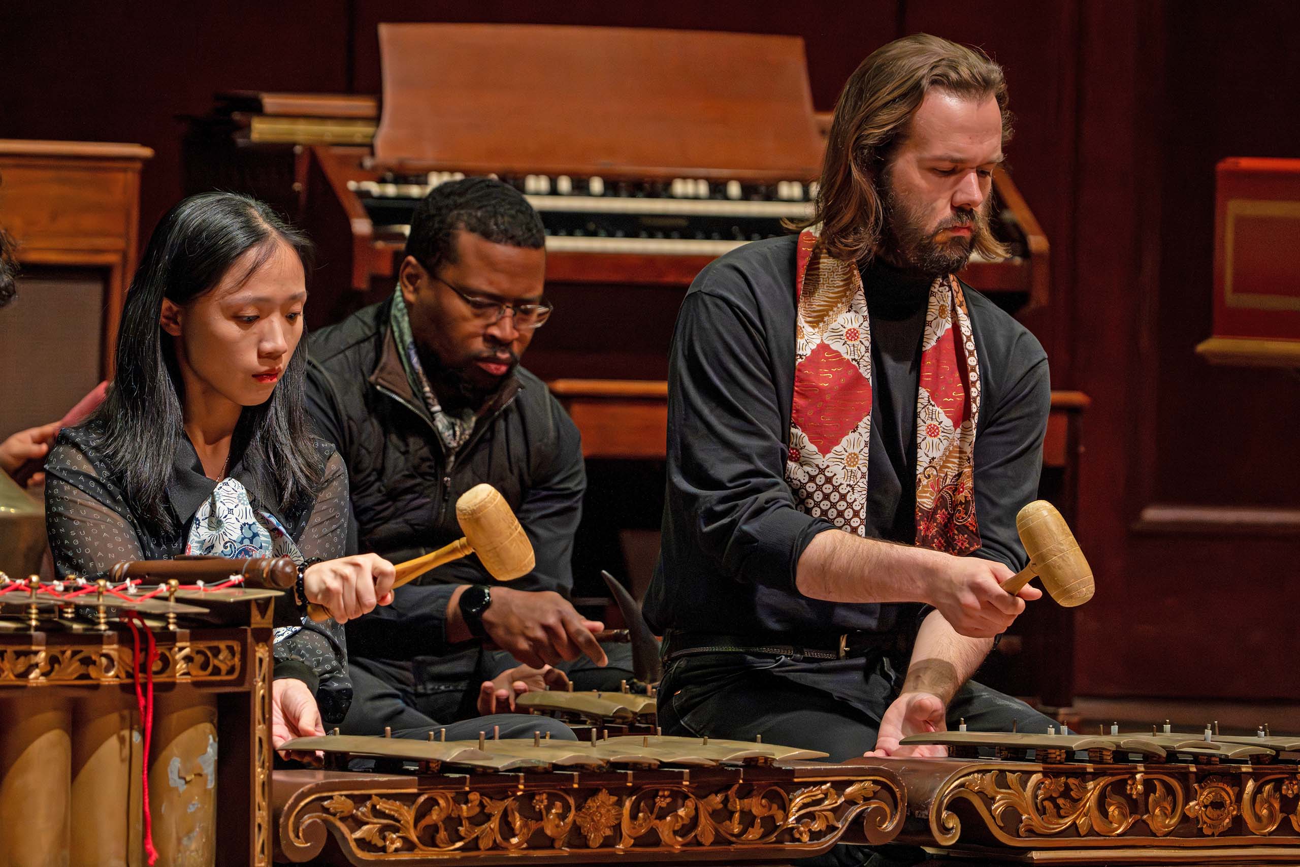 Three performers seated on a stage floor use mallets to play gamelan together in a concert.