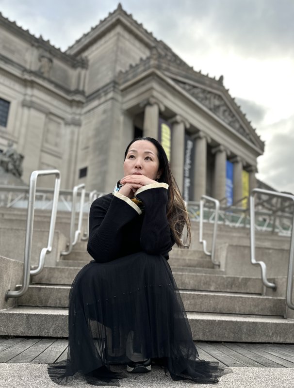 A photo of the speaker, Kanako Uzawa. She is a crouching outside of a large gray museum-like building, looking pensive up at the sky.