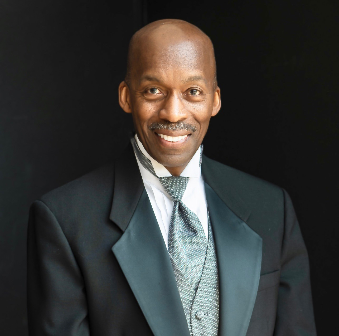 Formal studio portrait of Harvey Felder, attired in a black tuxedo coat with grey vest and tie.
