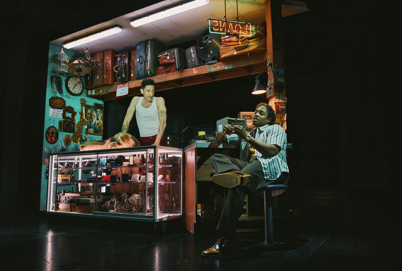 Two actors perform a scene on stage, set in a pawn shop. The lights inside a display case, flourescent overhead lights, and a neon sign that says LOANS stand out in an otherwise dark and shadowy set. 