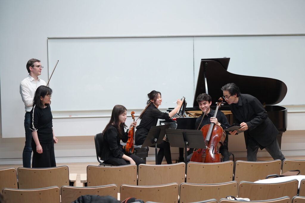 Six musicians engage in a group lesson in a classroom space, with a piano and string instruments. A large whiteboard is on the back wall, with rows of lecture seating in the lower foreground.