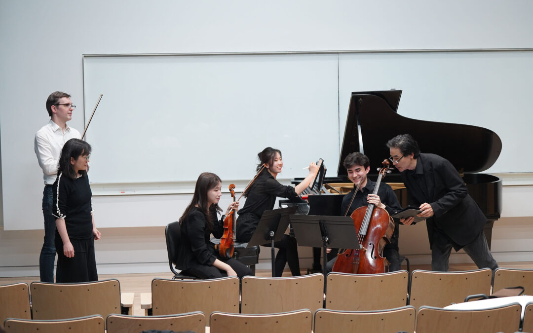 Six musicians engage in a group lesson in a classroom space, with a piano and string instruments. A large whiteboard is on the back wall, with rows of lecture seating in the lower foreground.