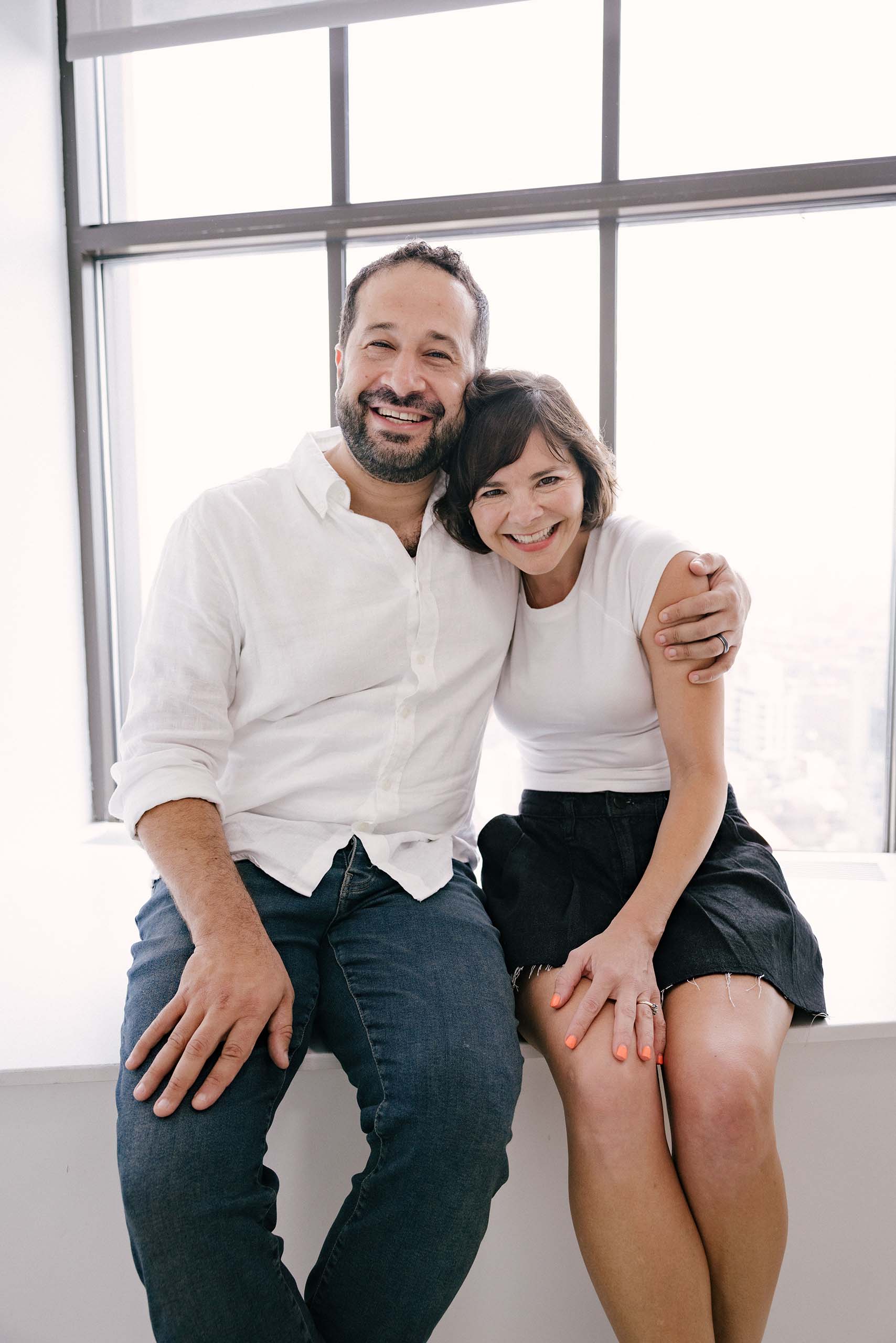 Portrait of Ian and Anne Eisendrath both in white shirts, with one arm each around the other, seated together with a large paned window in the background.