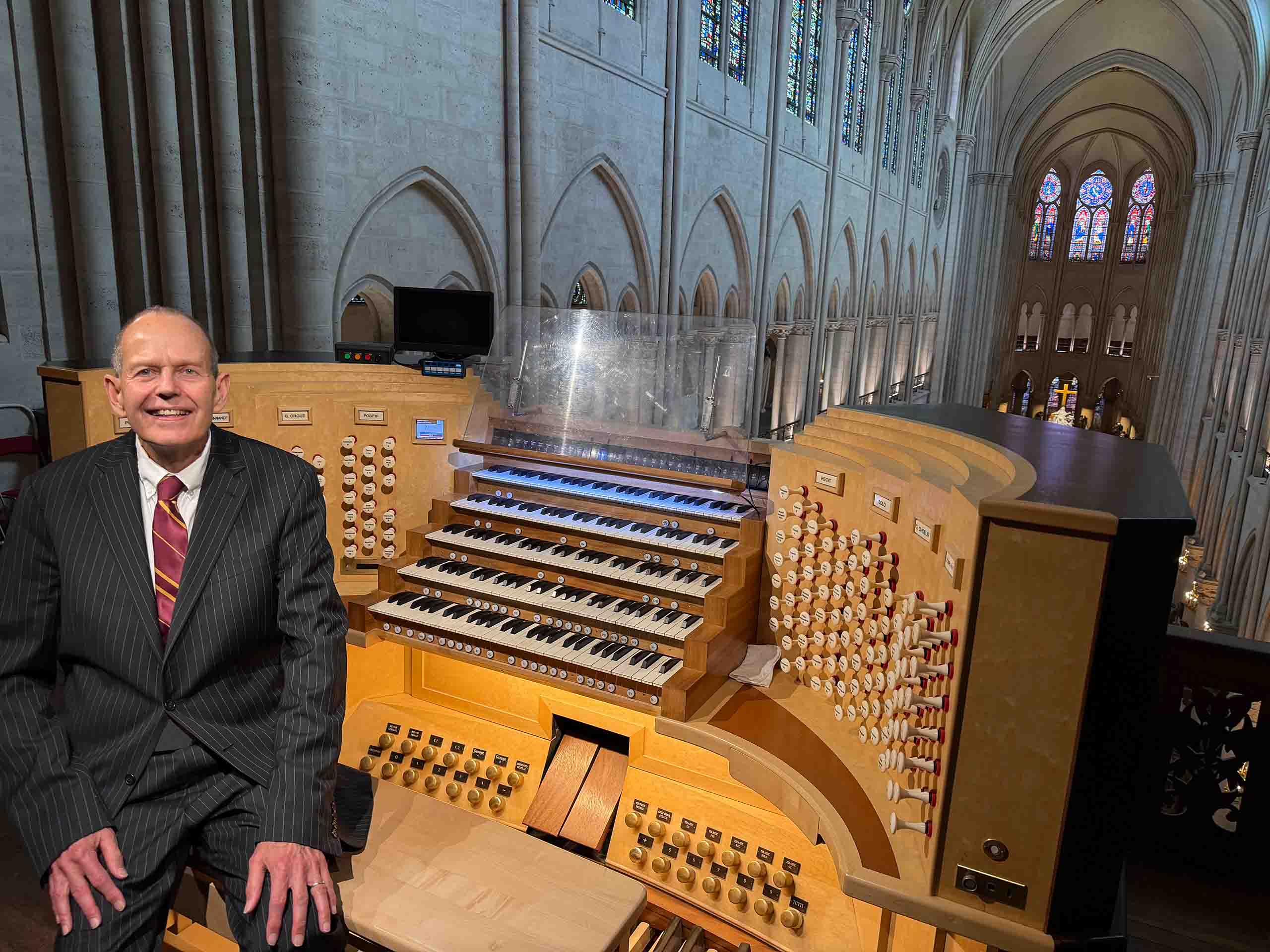 James Kibbe poses seated at an oak-hued organ console full of keys and buttons. Behind him in the background is the vast multi-story interior of Notre Dame cathedral and colorful stain glass windows.