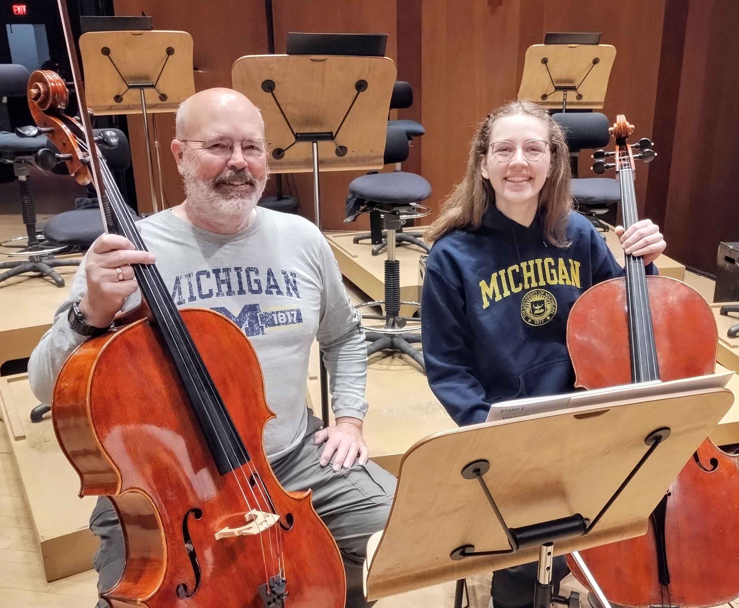 In a tiered rehearsal space with wooden music stands, Jeffrey Butler and Lauren Mathews are seated side-by-side, both wearing Michigan shirts and holding their cellos.