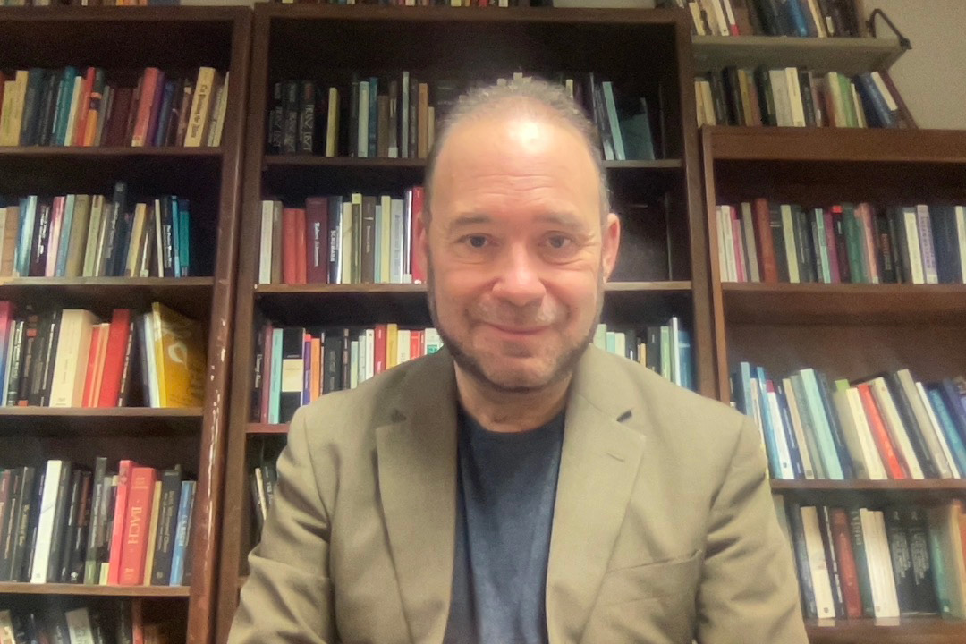 Casual headshot of Jeffrey Swinkin in a tan suitcoat, with a wall of full bookshelves in the background.