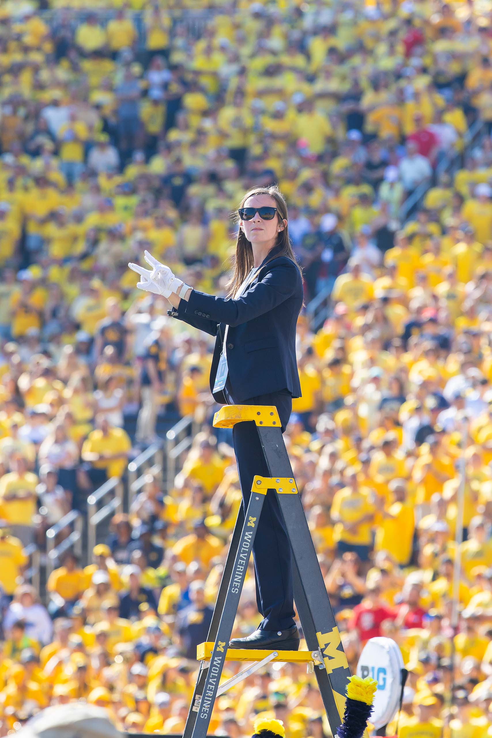 Wearing a blue suit, sunglasses, and white gloves, Jenn Hepp conducts from the top steps of a ladder. Behind her, the stadium crowd is full of fans wearing yellow.