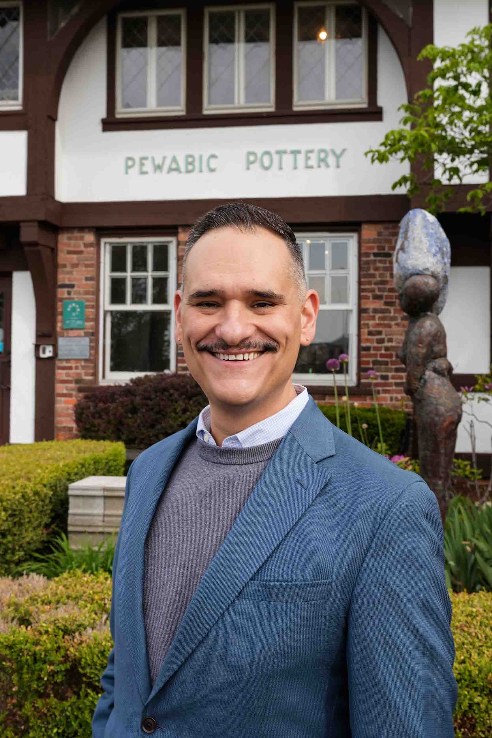 Jim Leija poses standing outdoors in a blue suitcoat and grey sweater, with the Pewabic Pottery sign on its stick style building in the background.