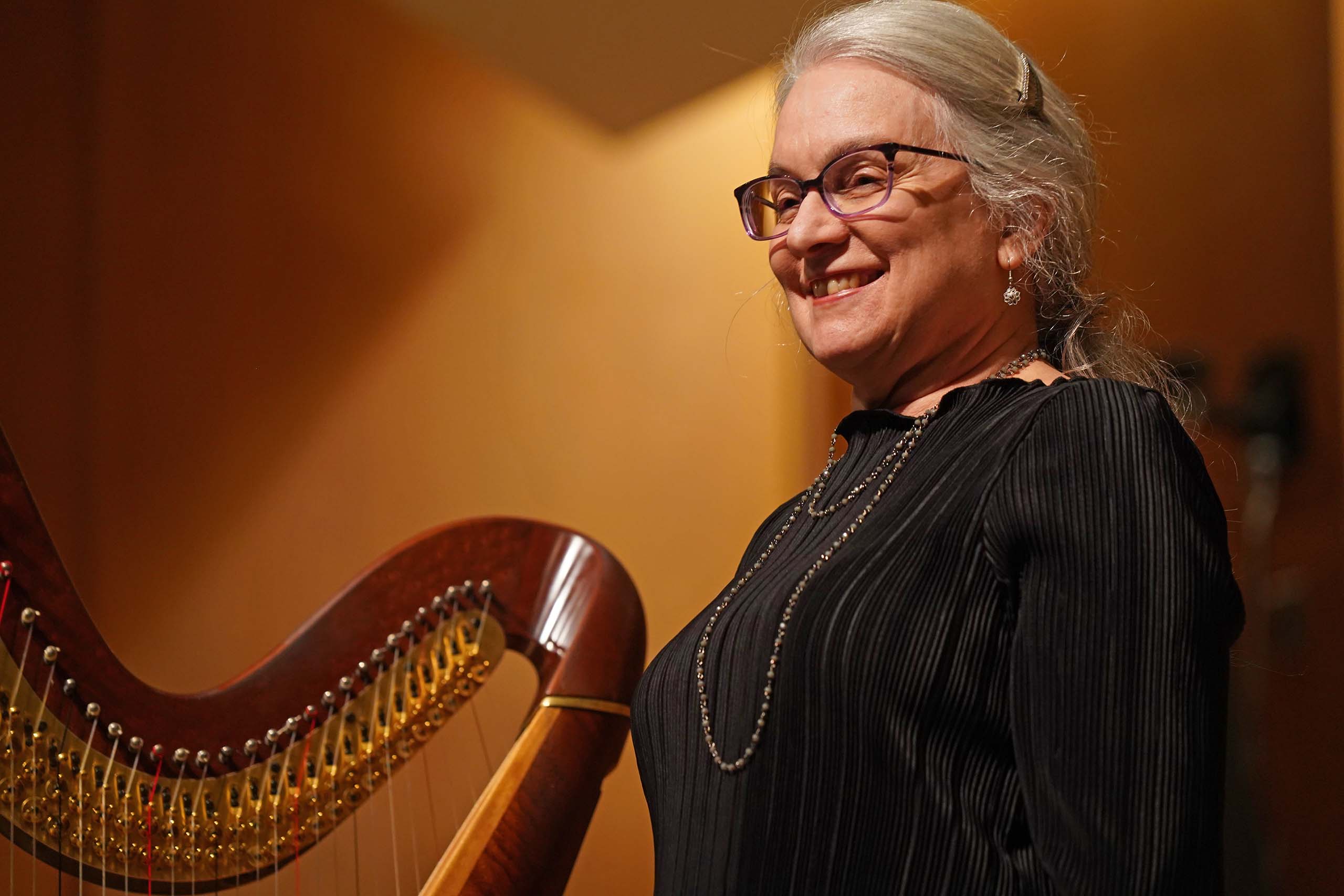 Joan Holland stands smiling under a stage light wearing black concert attire. The top of a harp is visible next to her.