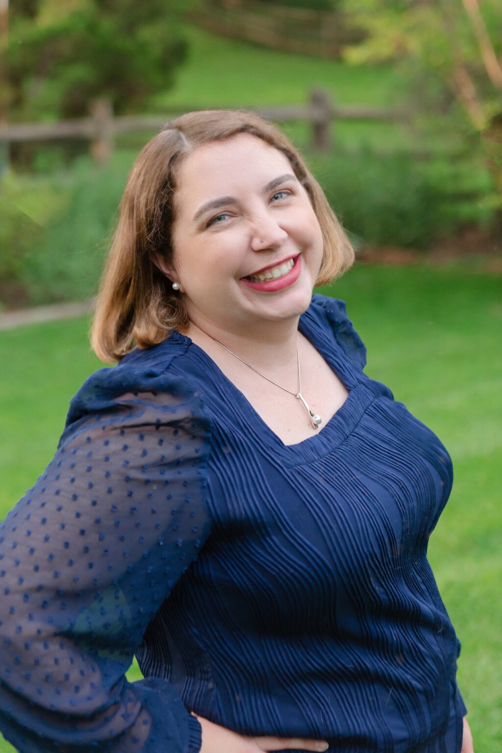 Portrait of Joanna Miller standing in a dark blue blouse, with green pastoral background.