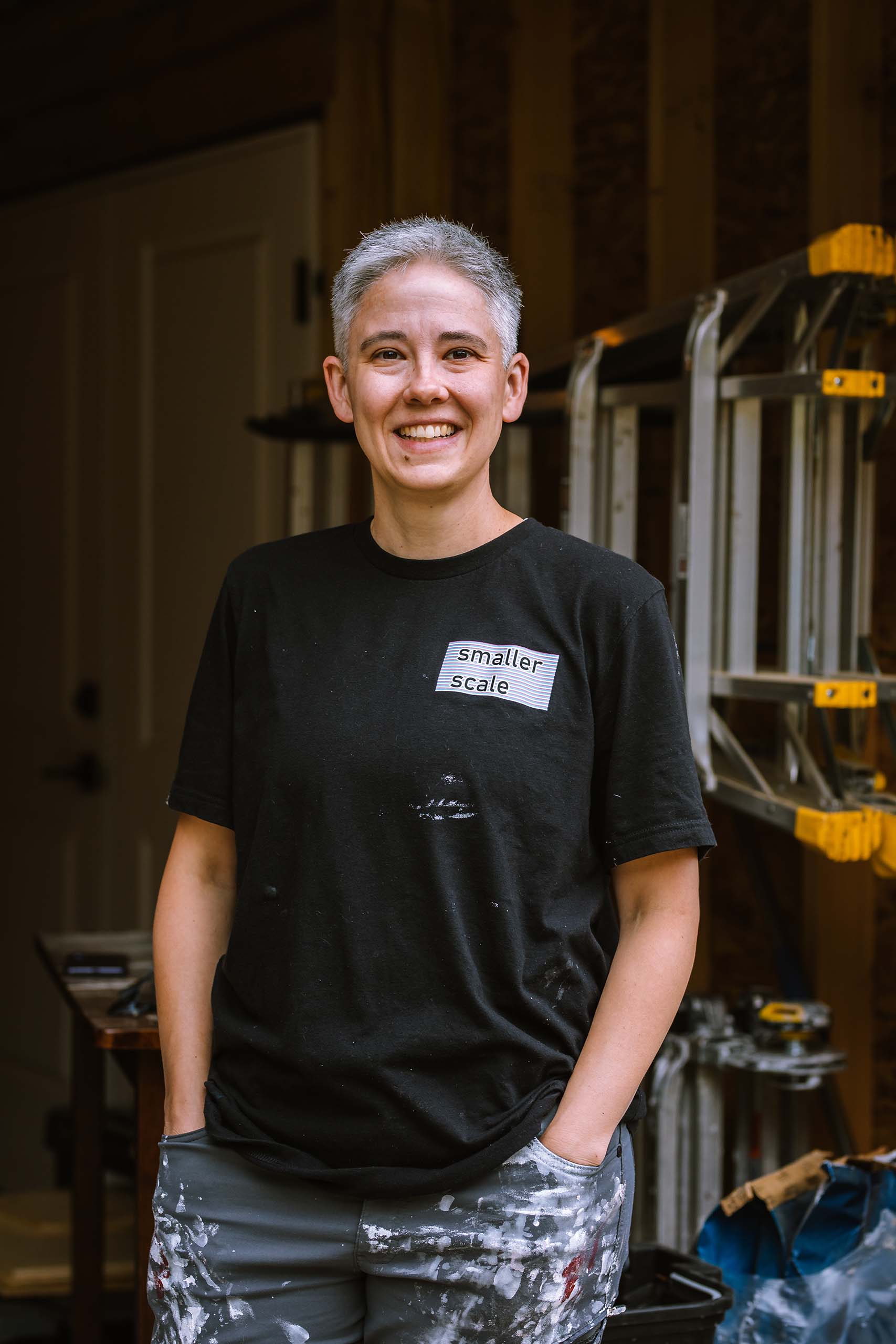 Johanna Schuster-Craig poses standing in a workshop space with ladders, attired casually with hands in her pockets.