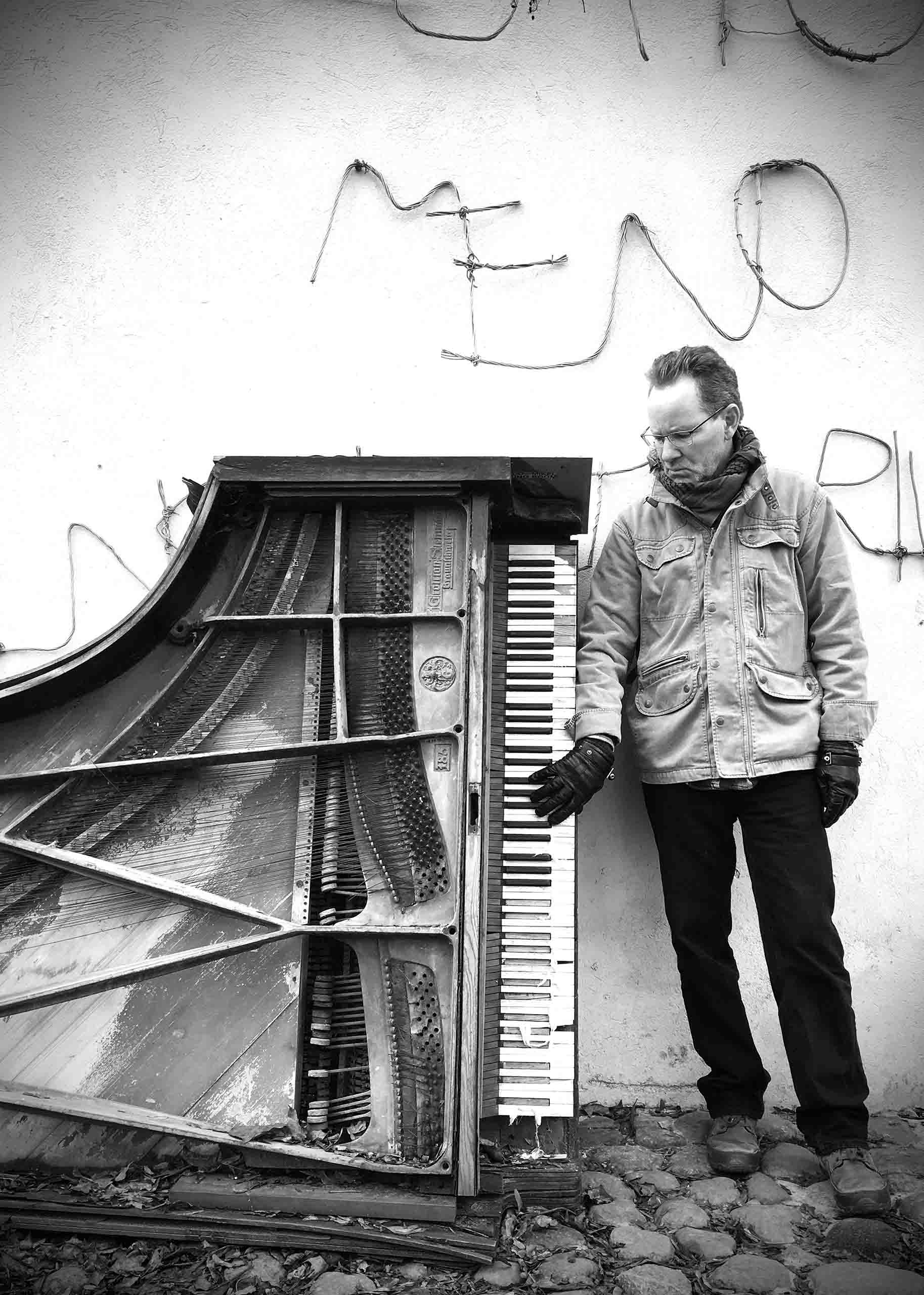 Greyscale portrait of John Mortensen standing next to a trashed baby grand piano with no legs, leaning on a concrete wall. He wears a winter coat and gloves.