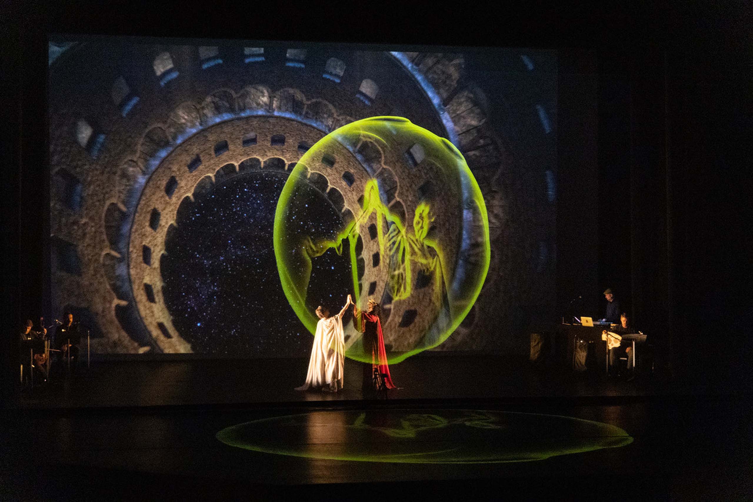 On a dark stage,a bright green projected orb shape hovers over two performers wearing long robes. In the background, musicians are seated on stage and a large projection of a futuristic space-scene fills the back wall.