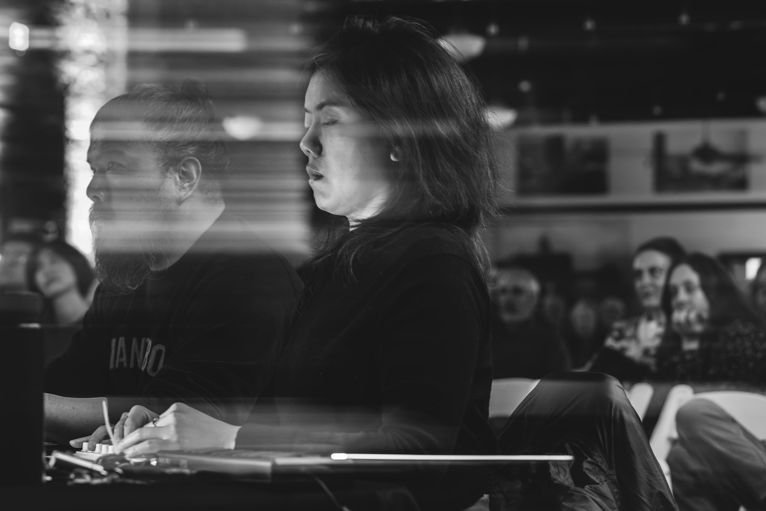 Greyscale photo of Julie Zhu seated and gazing down, using an electronic board with small dials. Other audience members are also seated in the background.