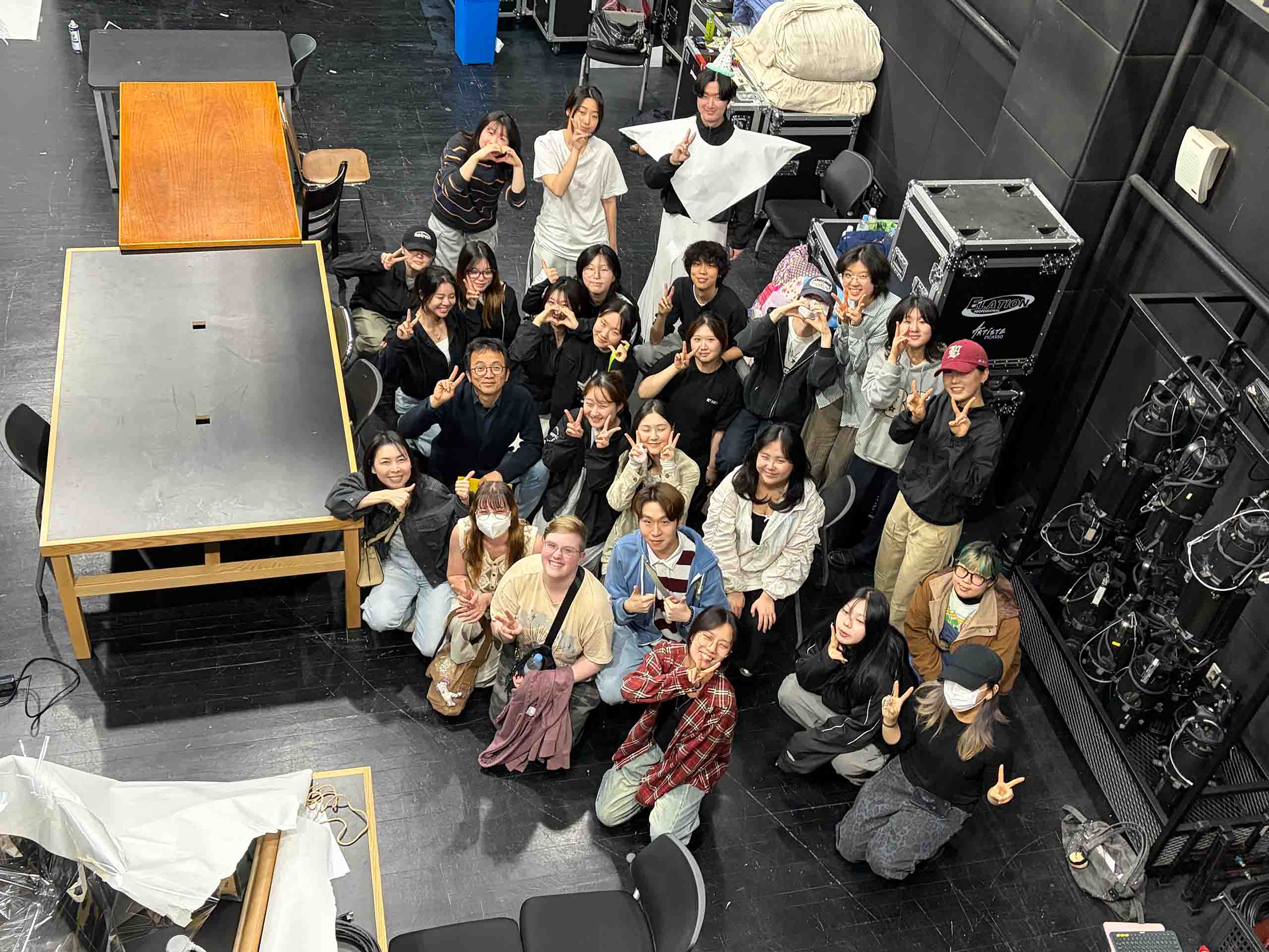About 27 people pose together in a backstage space, looking up a floor at the photographer above them. Some hold up peace signs or heart hands, and a few are wearing face masks.