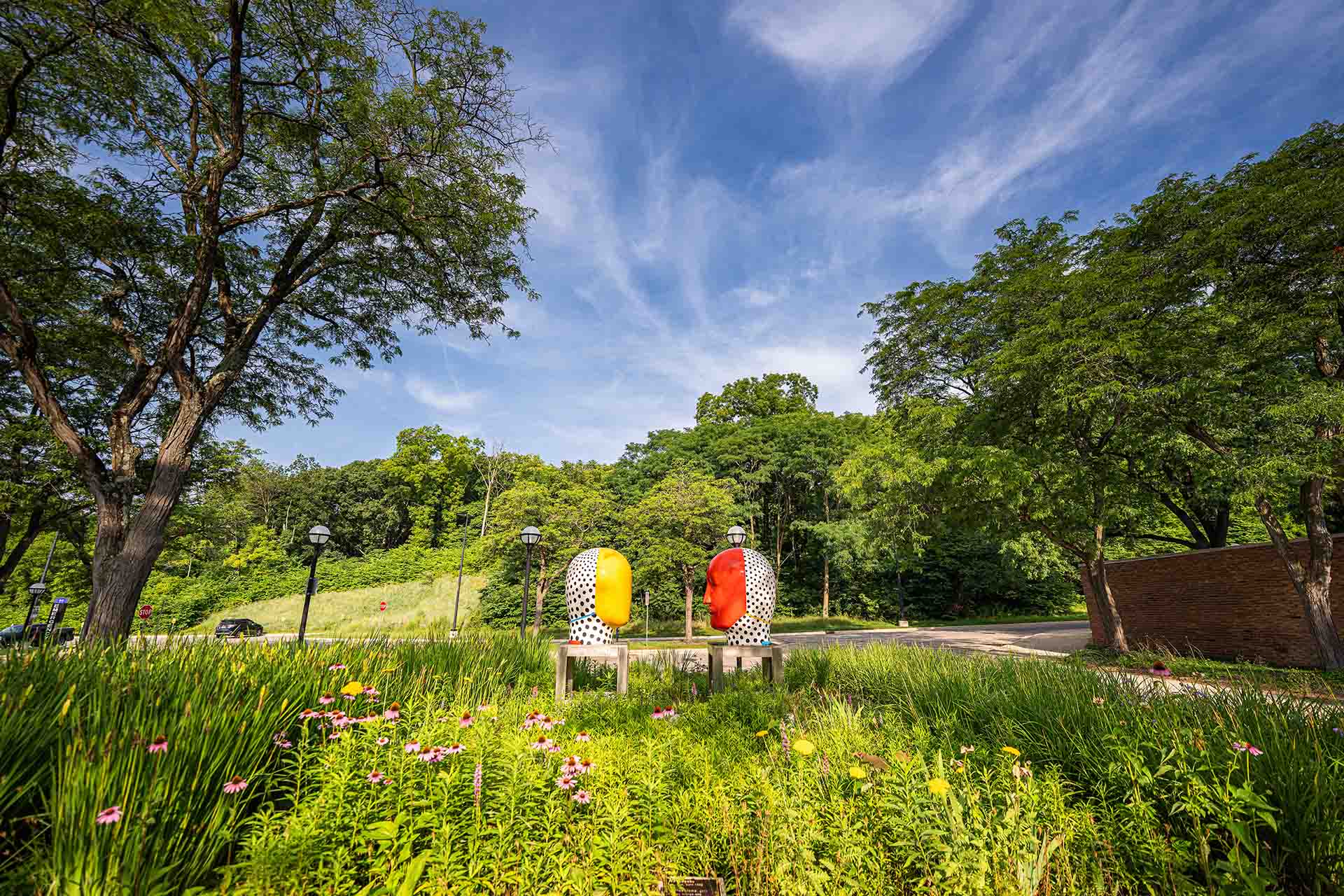 A campus sculpture of two large heads facing each other, one is yellow and the other is red. Summer flowers are in the foreground, and lush trees and a blue sky with wispy clouds surround the scene.