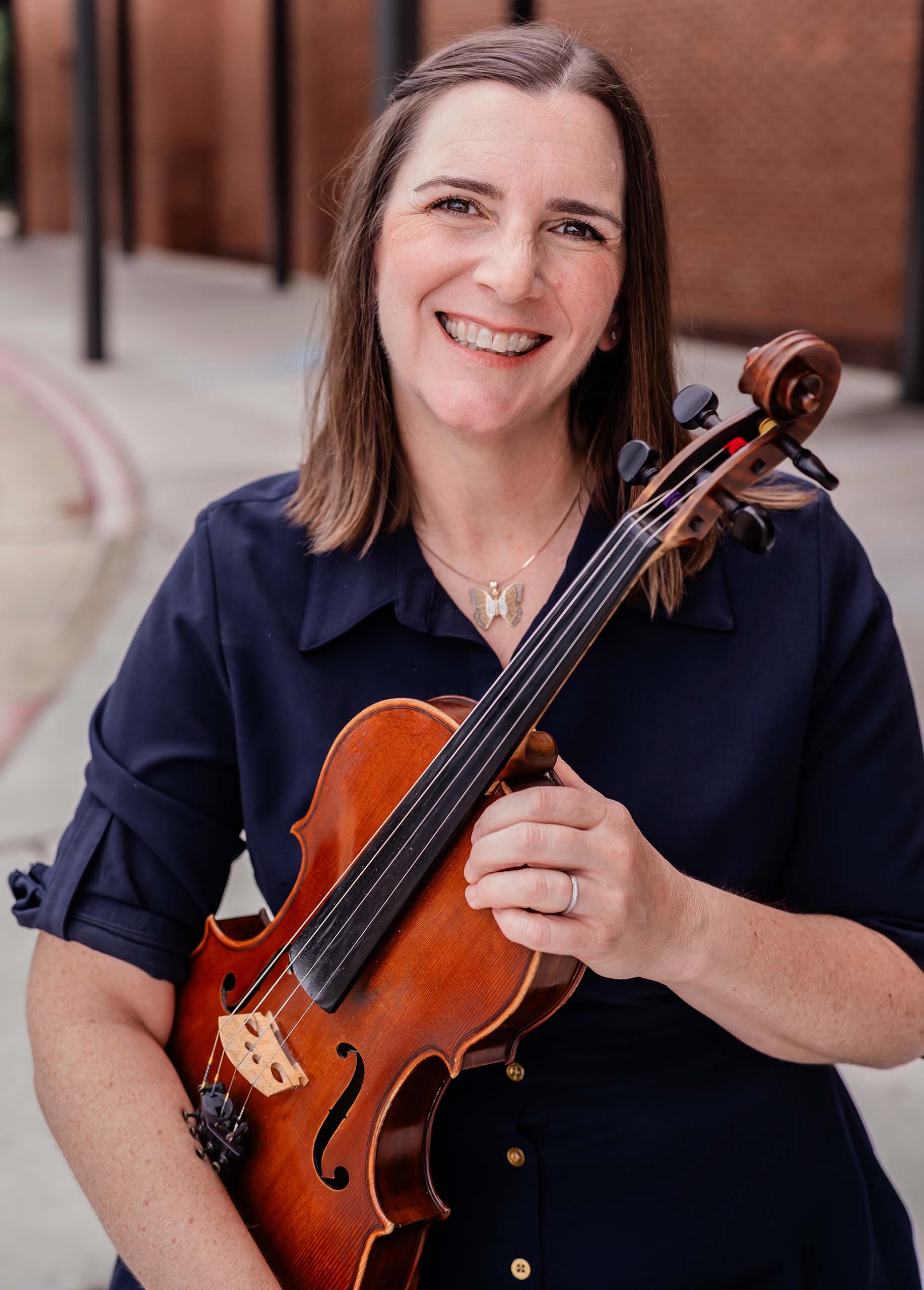 Studio portrait of Kate Francis standing on a street, holding a viola, in a dark blue dress with collar and buttons.