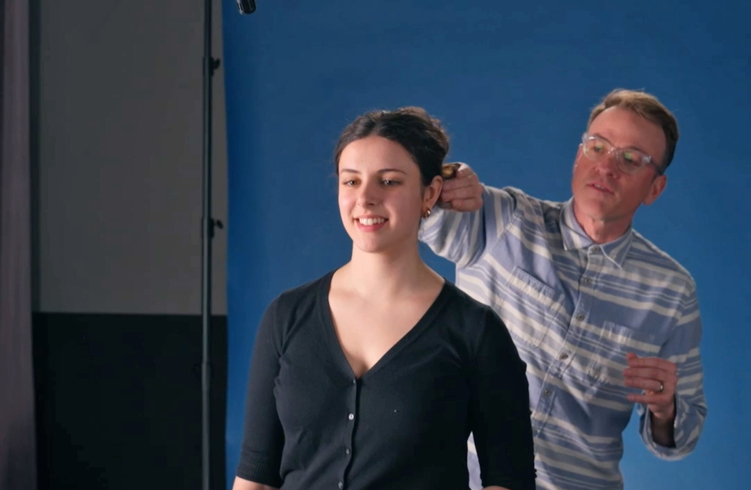 Standing in front of a blue recording screen, Katie Snowday smiles and holds still while Geoff Packard helps adjust something behind her ear.