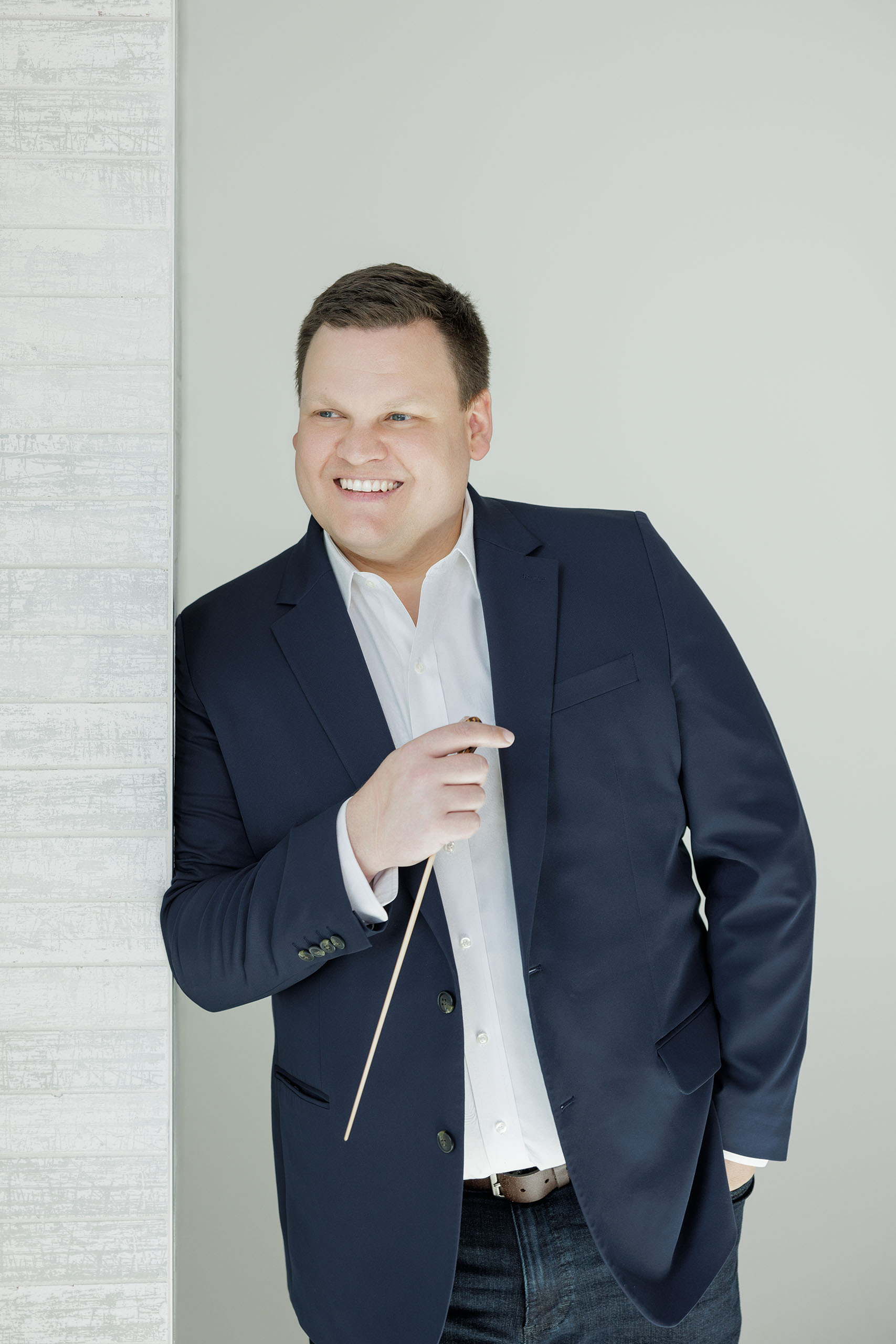 Formal portrait of Kenneth Sieloff holding a conductor's baton, leaning on an adjacent, light textured wall.