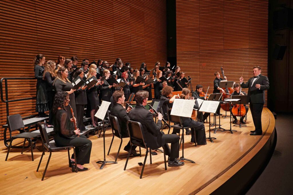 Kenneth Sieloff conducts a concert on an auditorium stage. He is conducting a choir of about 2 dozen singers performing with a ten piece orchestra.