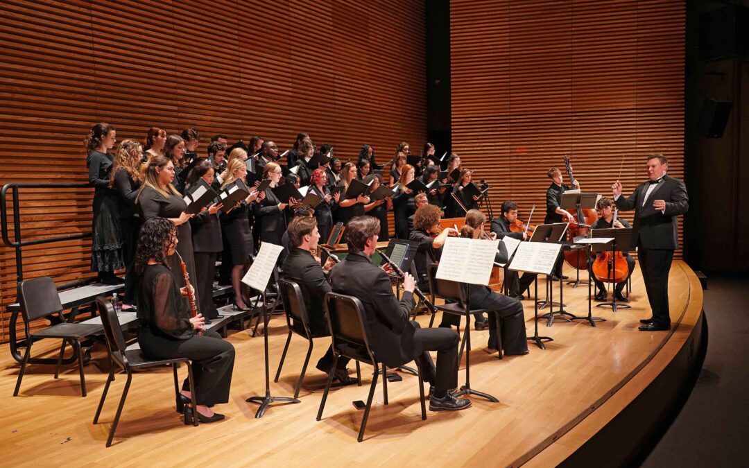 Kenneth Sieloff conducts a concert on an auditorium stage. He is conducting a choir of about 2 dozen singers performing with a ten piece orchestra.