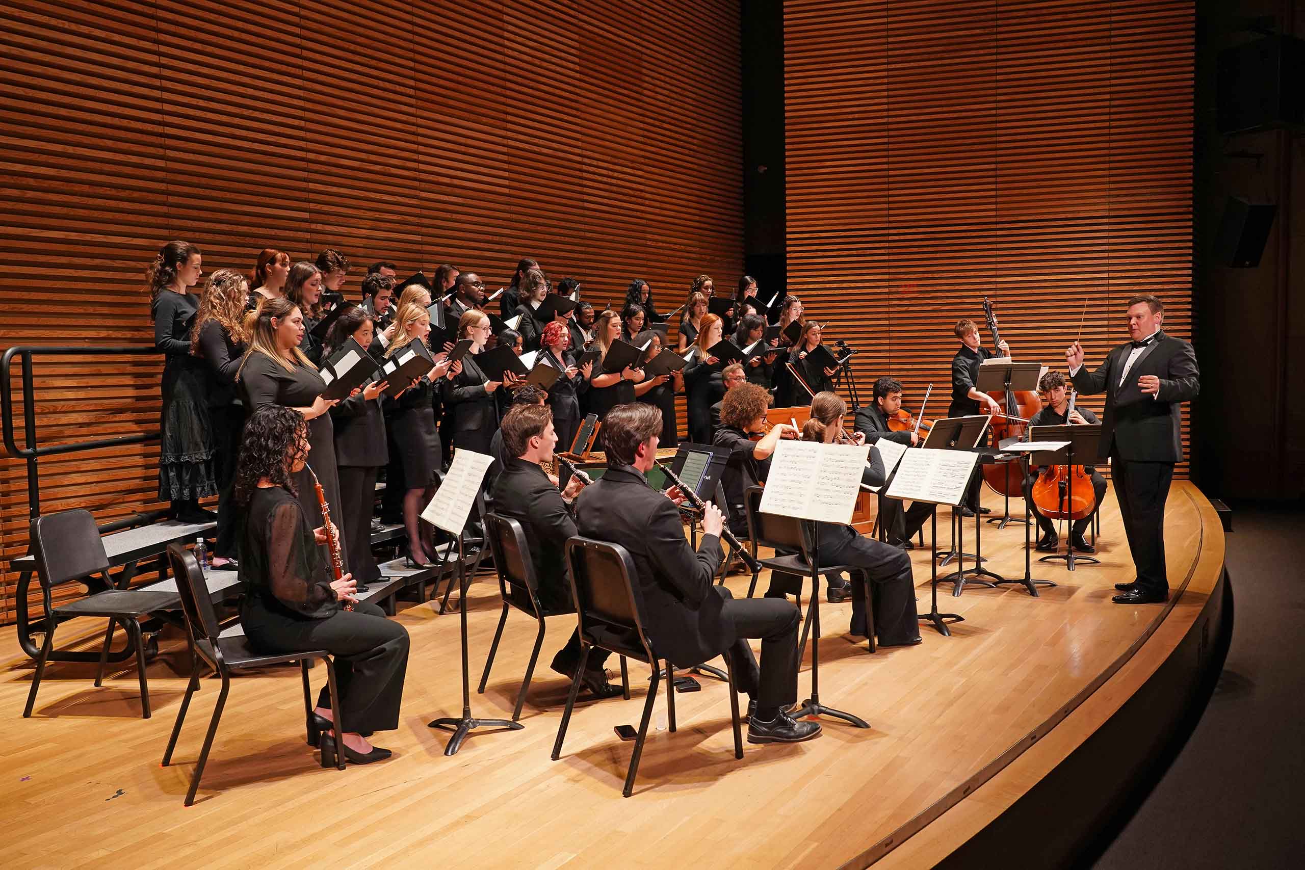 Kenneth Sieloff conducts a concert on an auditorium stage. He is conducting a choir of about 2 dozen singers performing with a ten piece orchestra.
