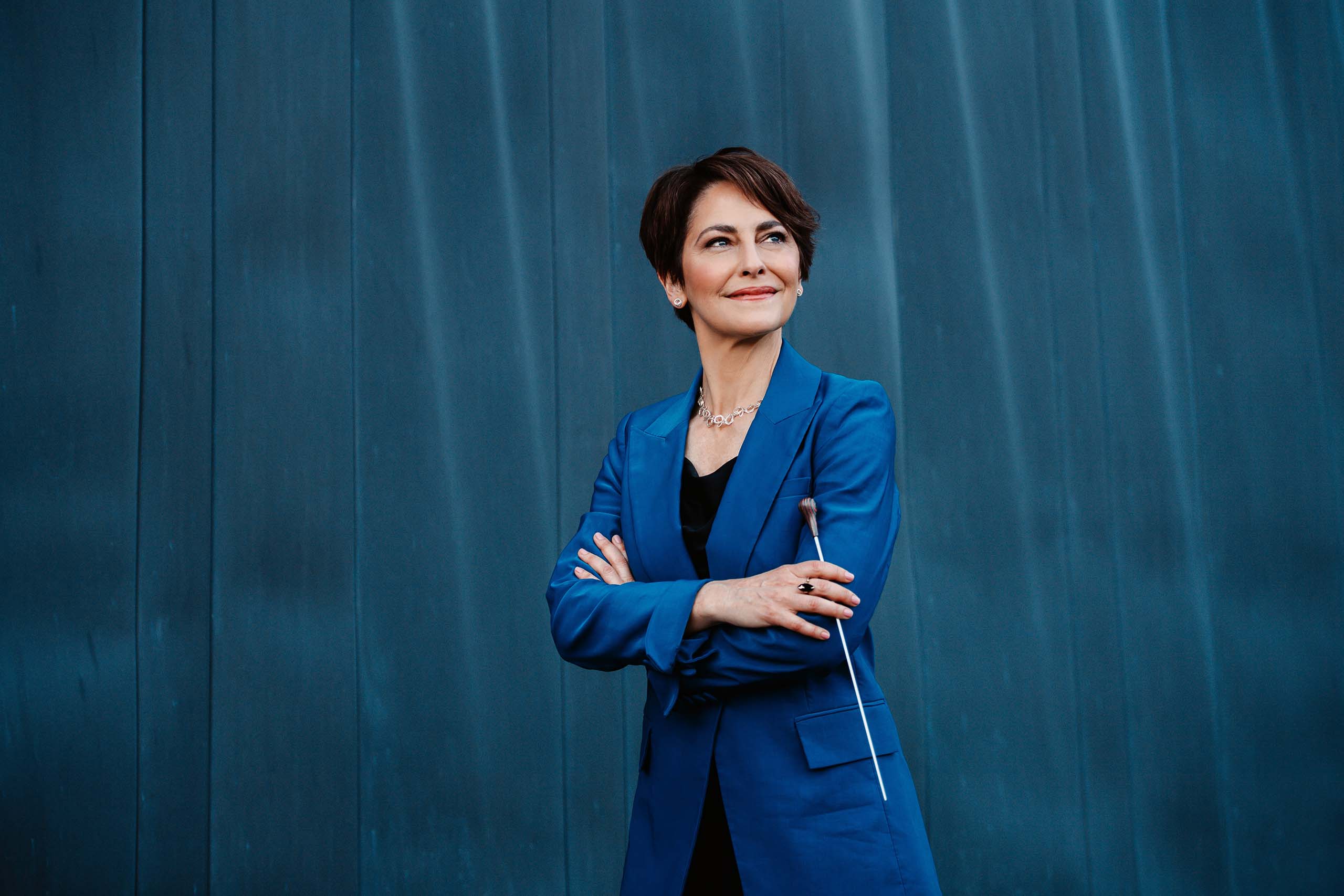Standing portrait of Laura Jackson with arms folded, holding a conductor's baton. She wears a blue suitcoat, and a teal textured wall fills the background.