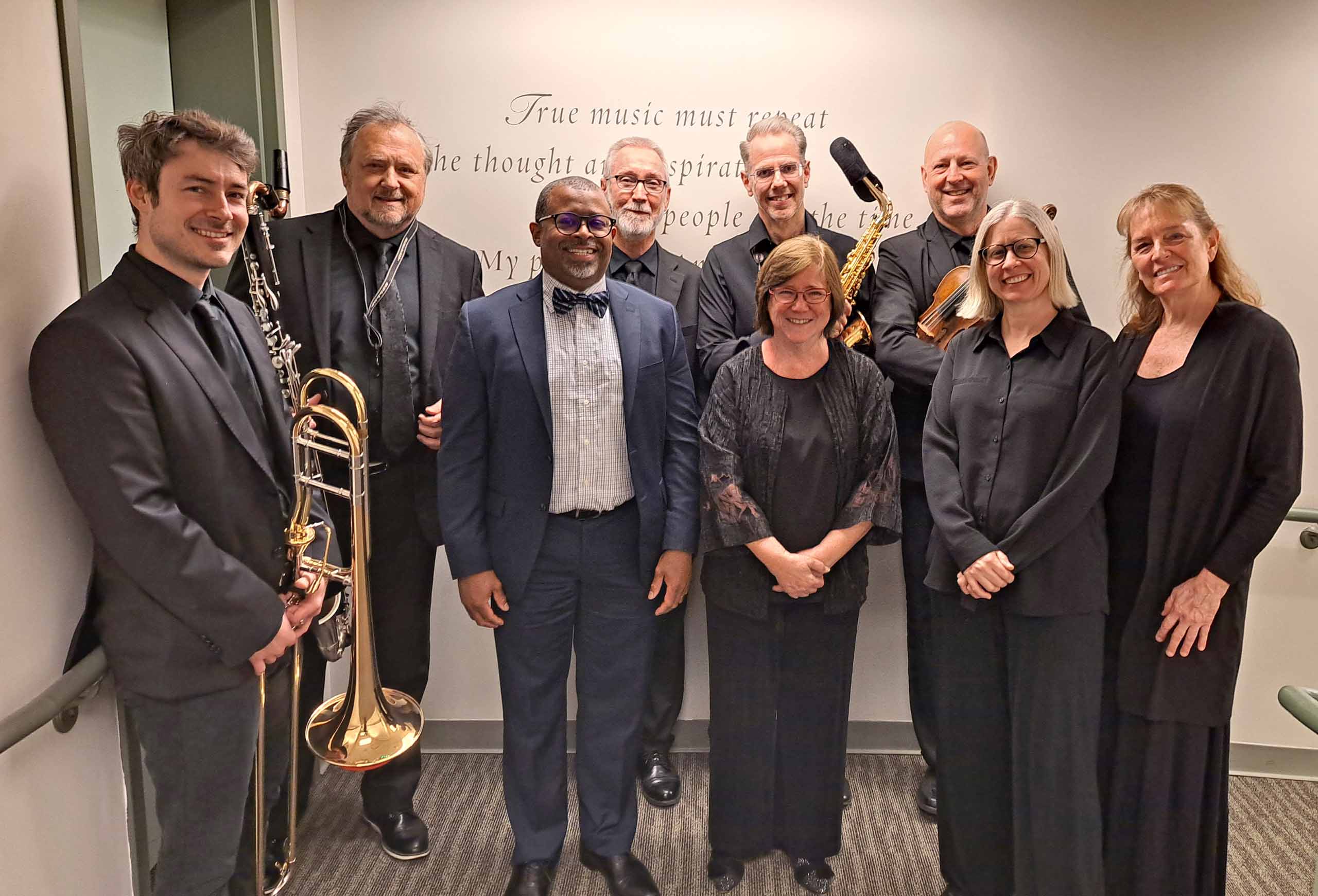 Nine people pose standing together in two rows, wearing formal attire. Four of them hold instruments.