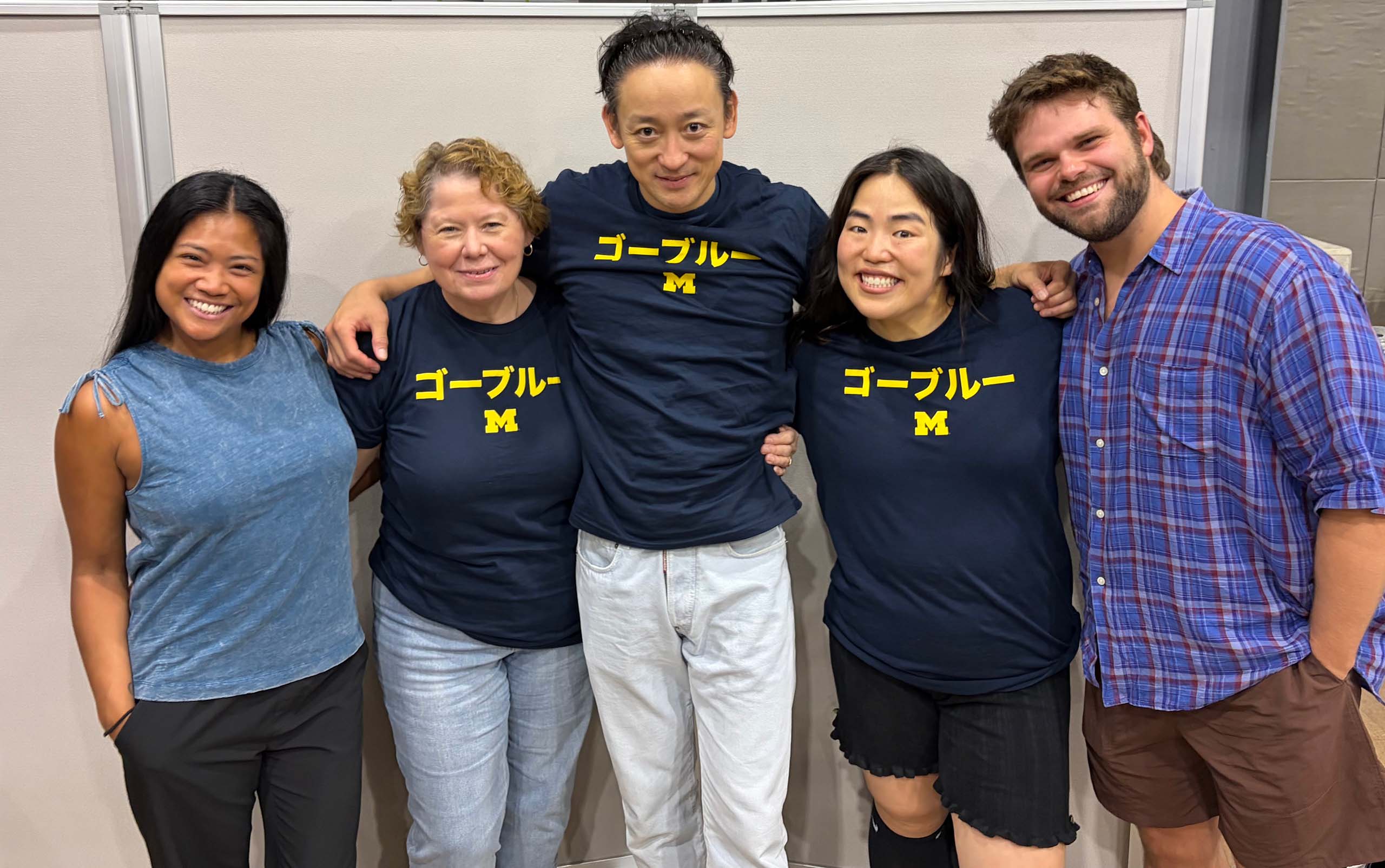MT alums Shea Renne Griffin Binnicker Catherine Walker Koji Yamamoto Yuriyan Retriever t-shirts Go Blue in Japanese Five people pose standing together, with arms around each other, attired casually. The three in the middle all wear blue t-shirts with a yellow block M and text in Japanese.