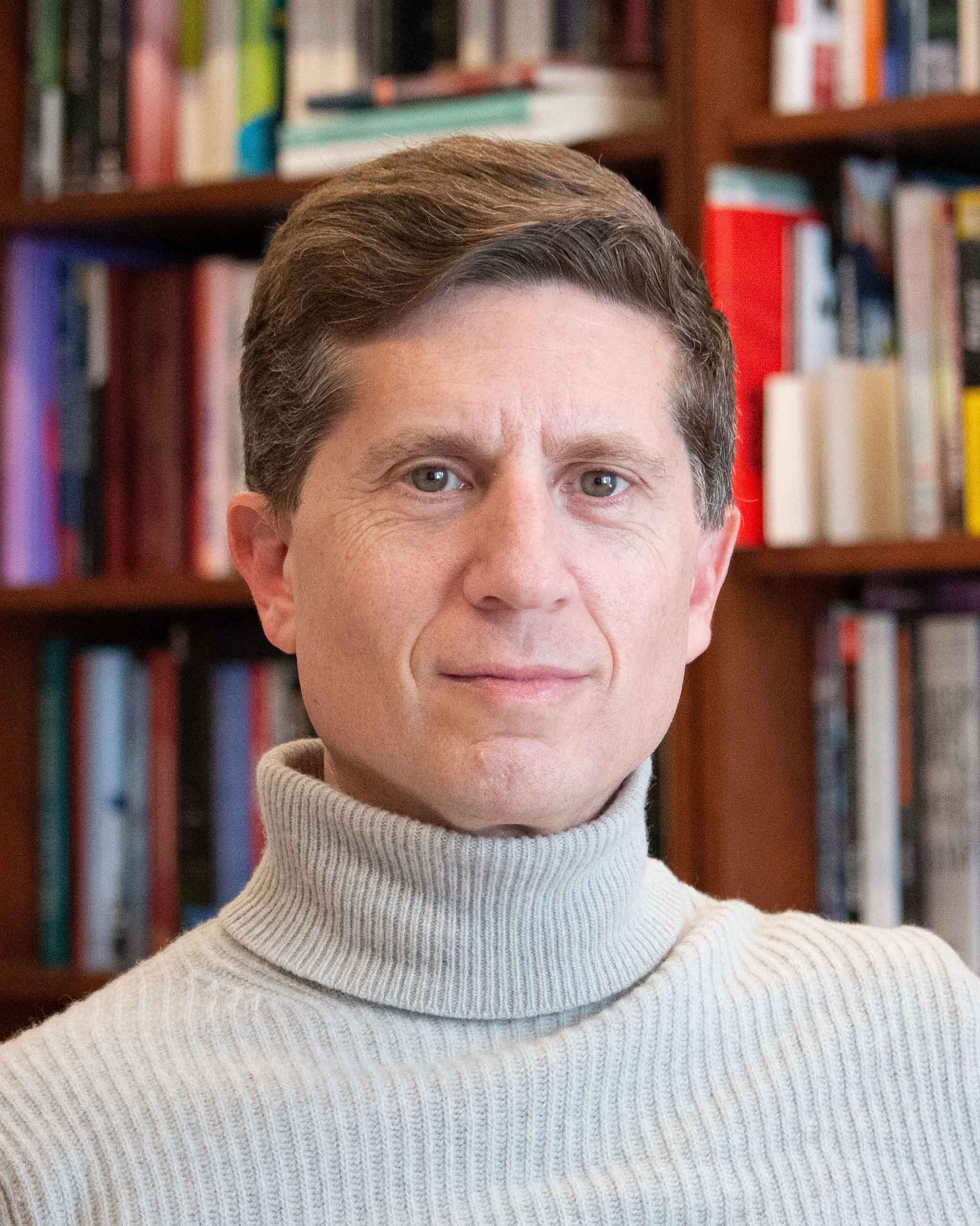 Headshot of Mark Katz wearing a grey turtleneck, with shelves of books in the background.