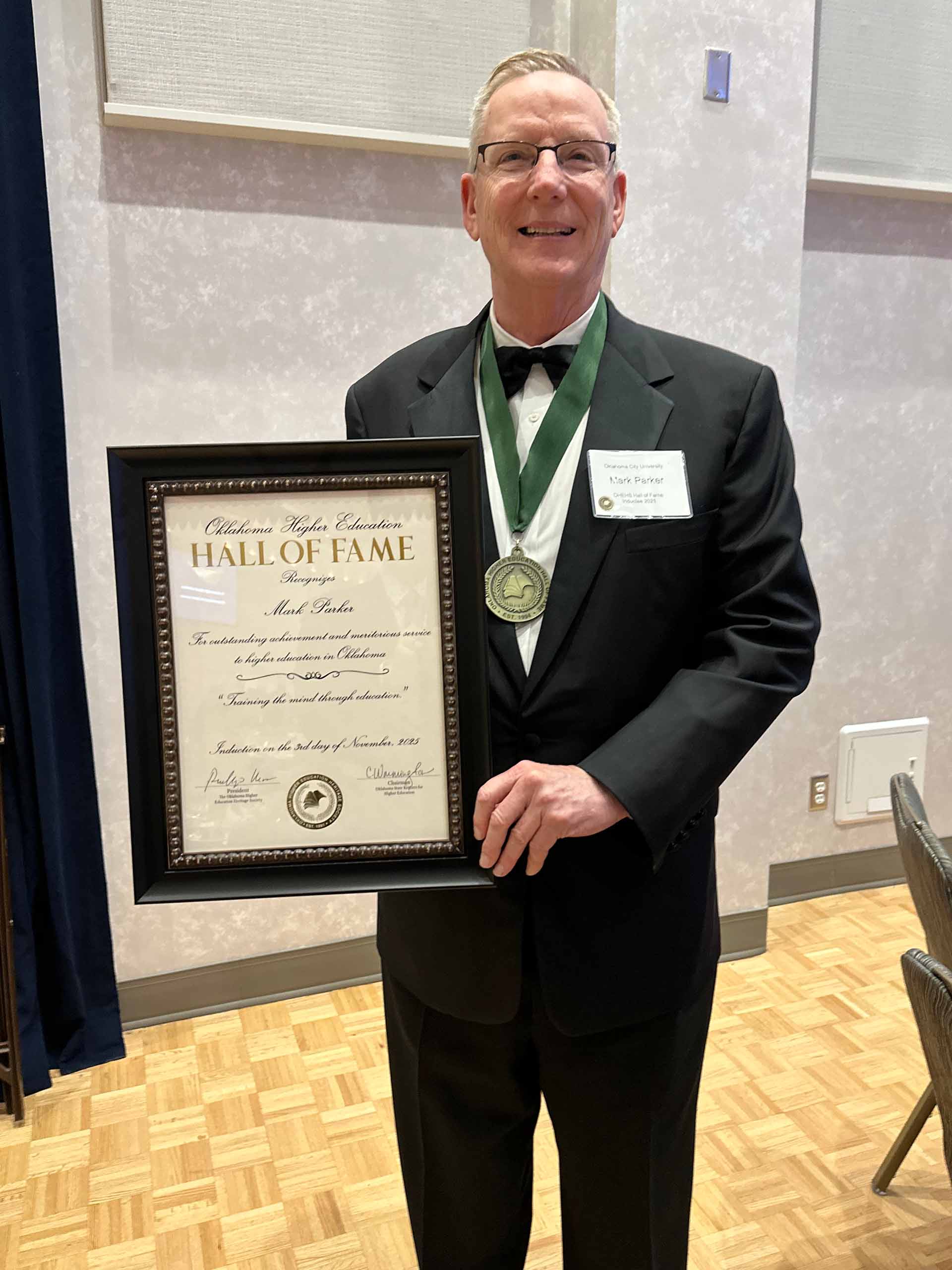 Mark Parker poses standing, wearing a ribbon medal and holding up a framed Hall of Fame certificate. He wears a black tuxedo and a name tag.