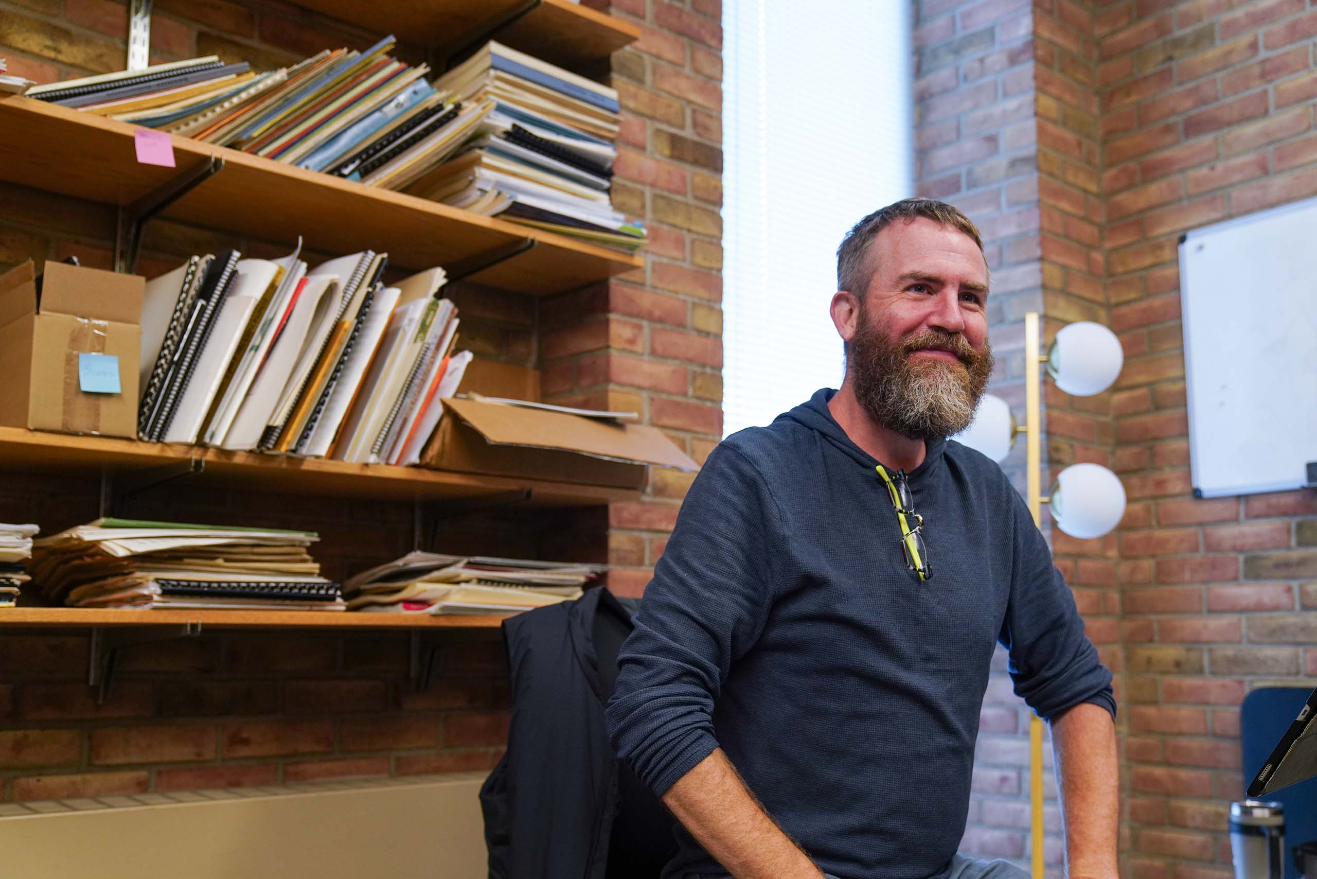 Candid photo of Matt Albert seated in an office space with brick walls, a window and a small white board. Behind him, several shelves are filled with stacks of sheet music, which have thin spiral bindings and folders.