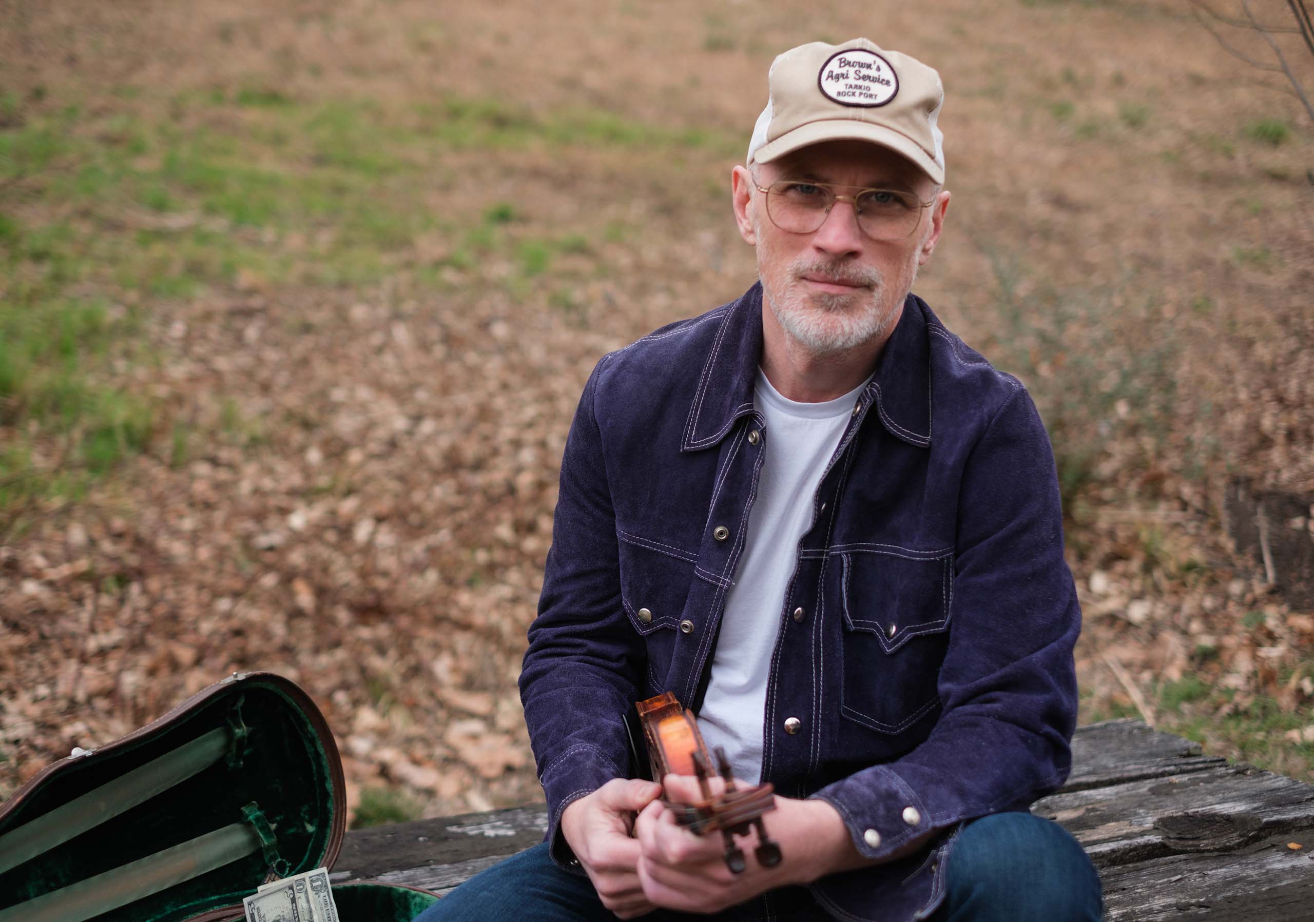 Portrait of Matt Combs holding his violin, seated outdoors next to his open violin case with dollars in it. He wears a jean jacket and ball cap. Fall leaves on the grass in the background.