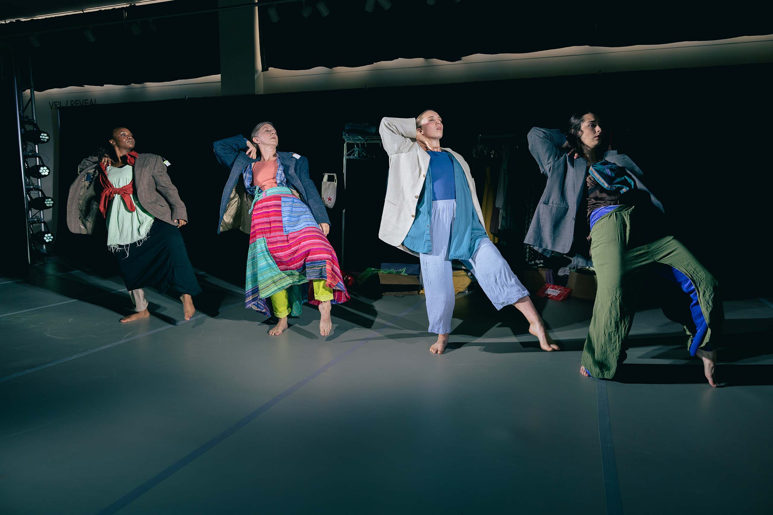 In a darkened studio space, four dancers stand in a line side-by-side, posed alike with one toe pointed and the opposite elbow bent and raised up high. They are costumed each differently in layers of loose fabrics and all wearing a cloth suit coat.