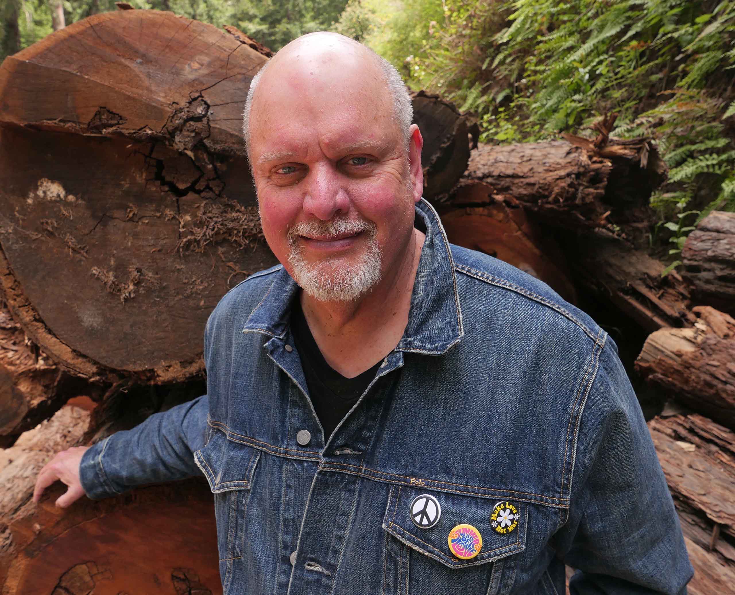 Headshot of Michael Daugherty standing outdoors in nature in front of fallen tree logs, attired in a blue jean jacket with 3 peace buttons.