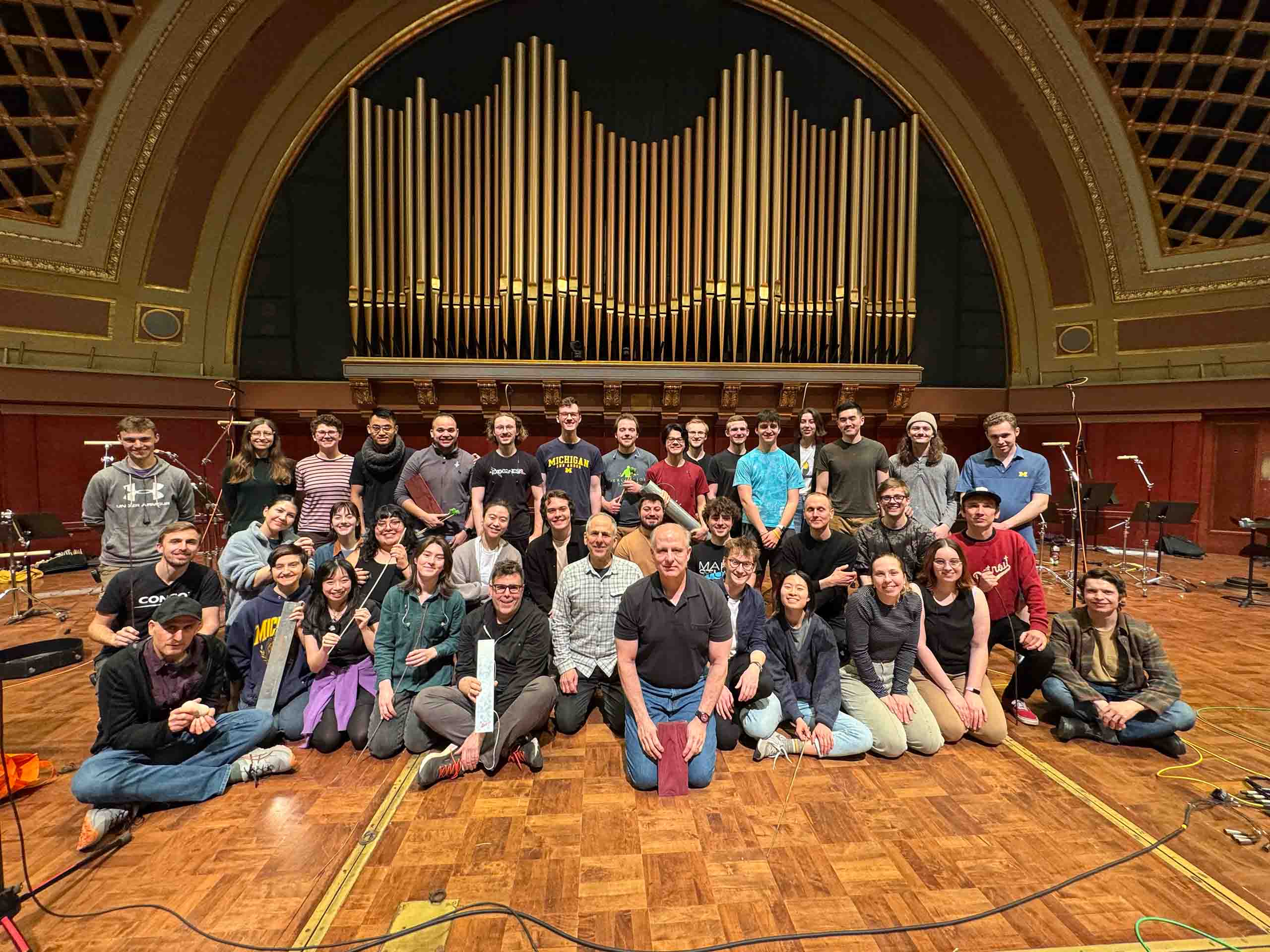 About 40 people pose as a group on the stage of Hill Auditorium in several rows, with some seated in front. Wires and microphones for recording surround them.