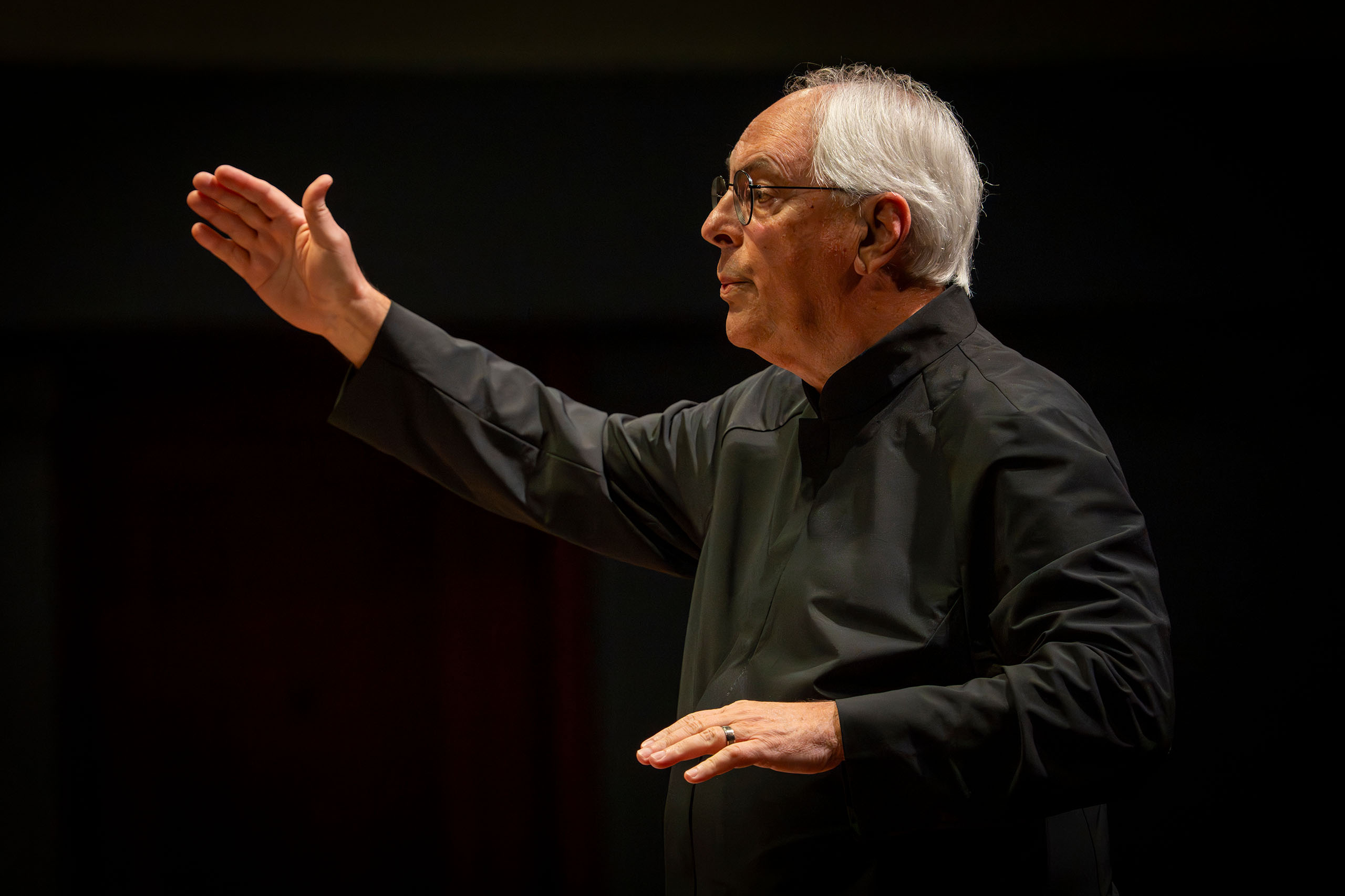 Michael Haitcock conducts in a dark performance space with no baton, attired in a black collared shirt.
