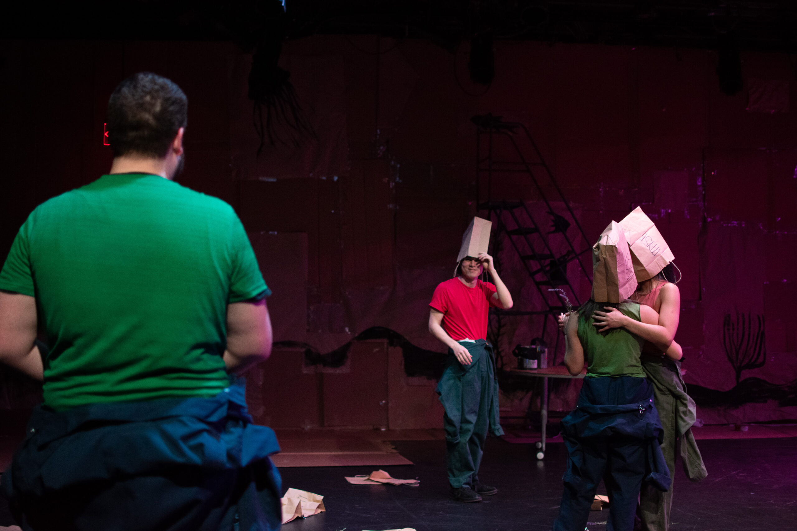 Three performers stand on a stage set with a dark, abstract background. Two are hugging while wearing paper bags on their heads; the third, in a red shirt, looks on with a playful gesture, also wearing a paper bag hat.