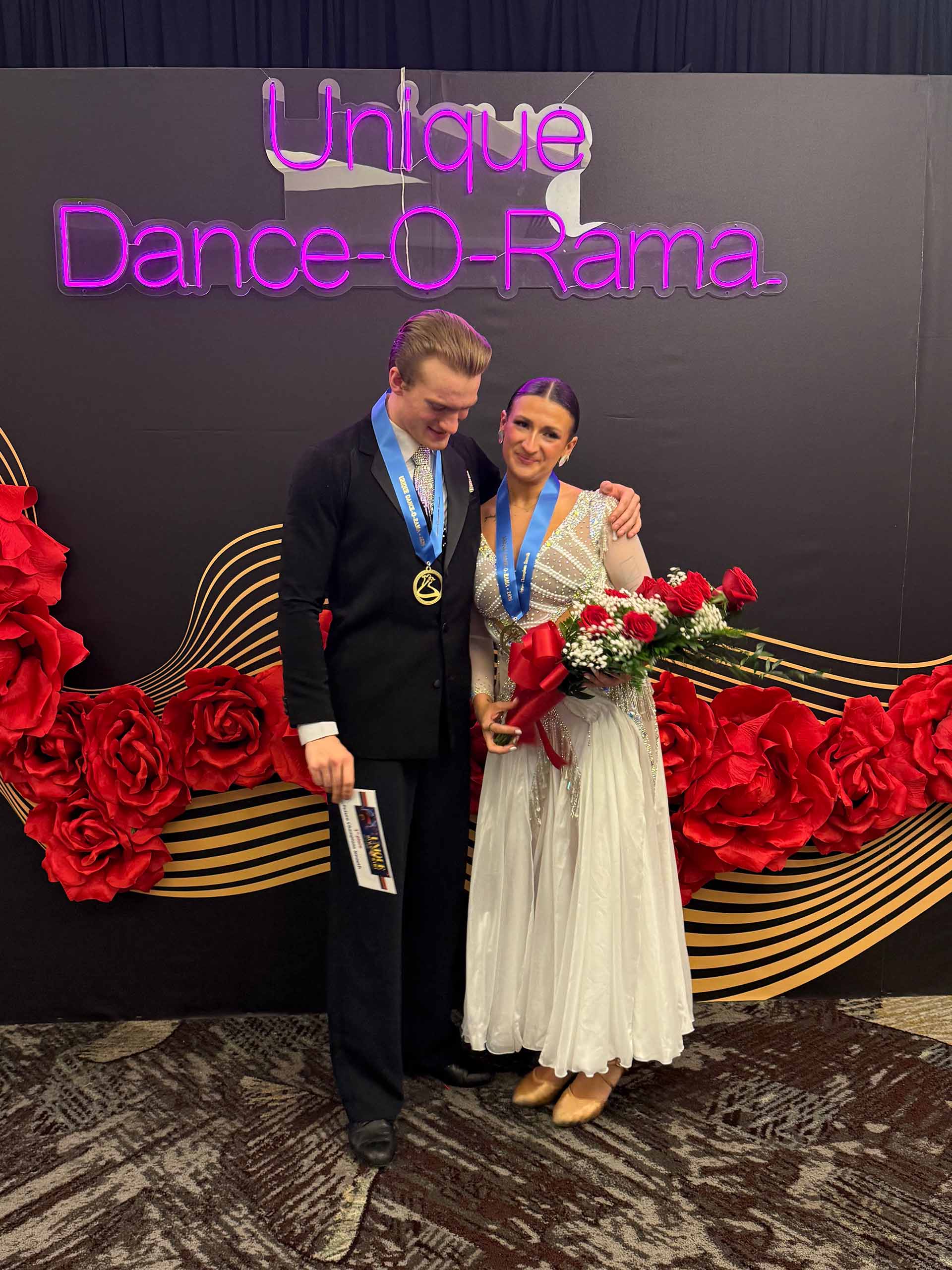 A dance couple in formal attire poses wearing medals with blue ribbons around their necks; one holds a bouquet of roses. In the background are large red roses and a neon sign that reads "Unique Dance-O-Rama"
