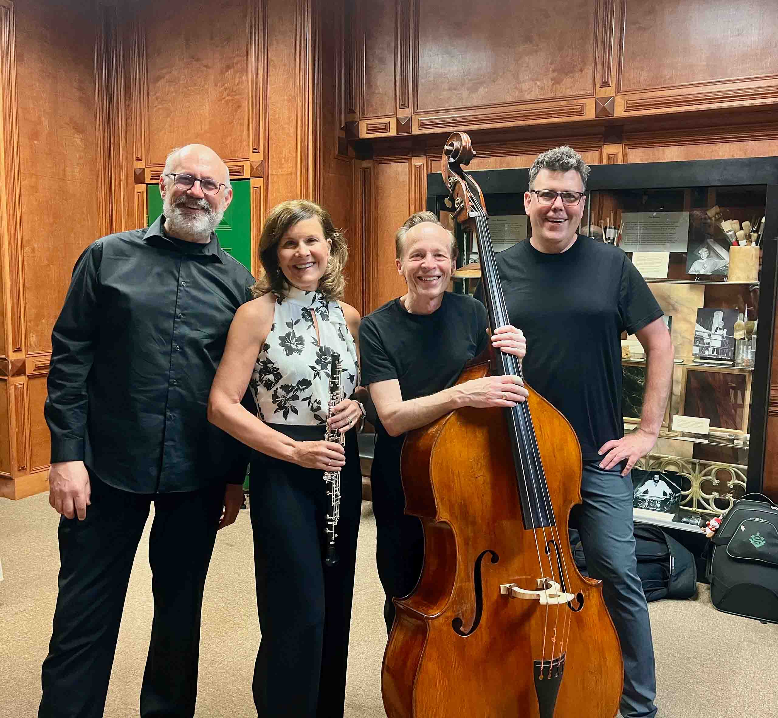 Four people pose standing together - one holds an oboe, and one holds a double bass. In the background we see display cases and wood-paneled walls.