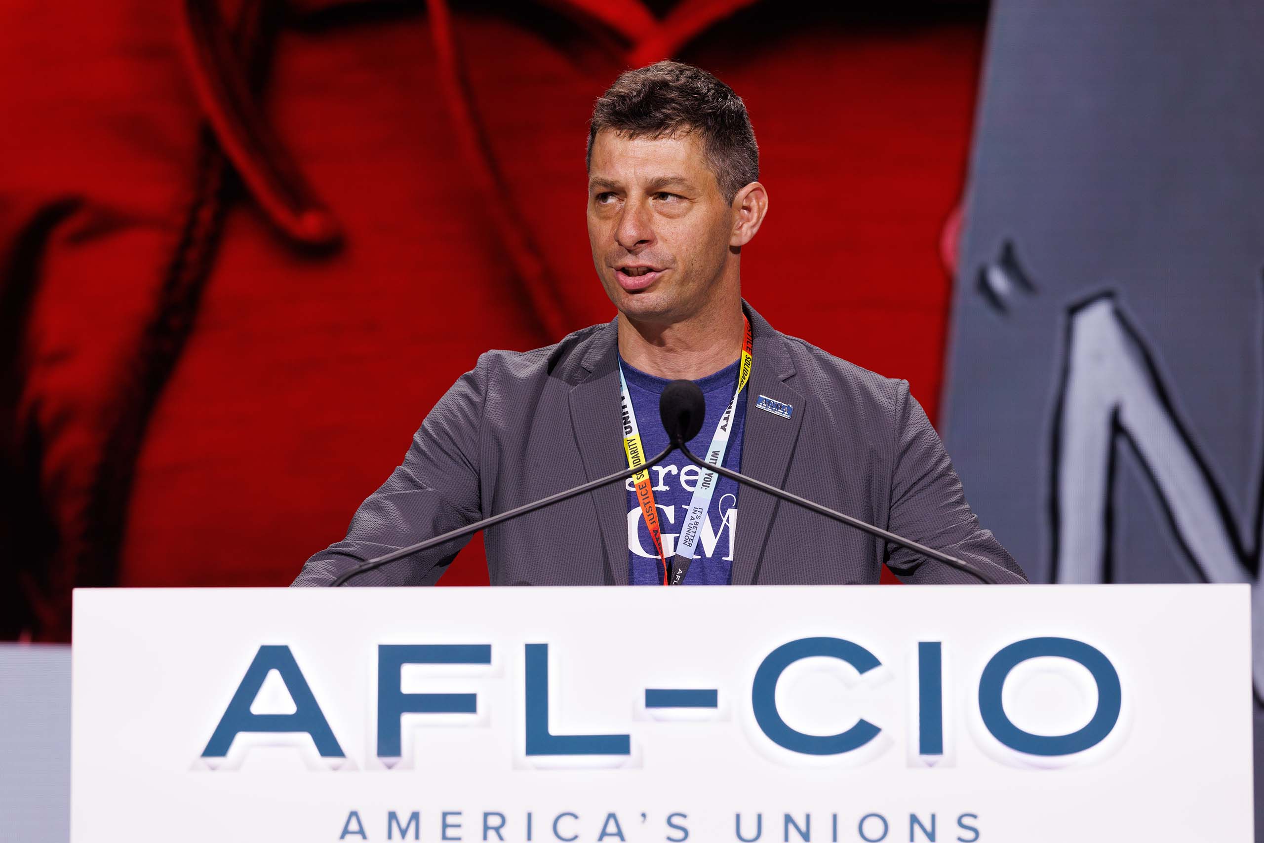 Ned Hanlon speaks behind a podium that says AFL-CIO America's Unions. He wears a gray suitcoat and a conference lanyard.