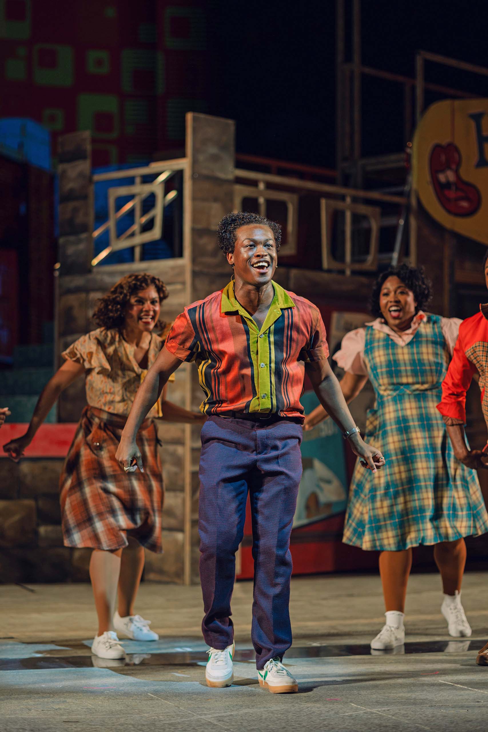 Performing on stage, Nicholas A. Wilkinson and two castmates sing and dance, costumed in mid-20th century youthful attire.