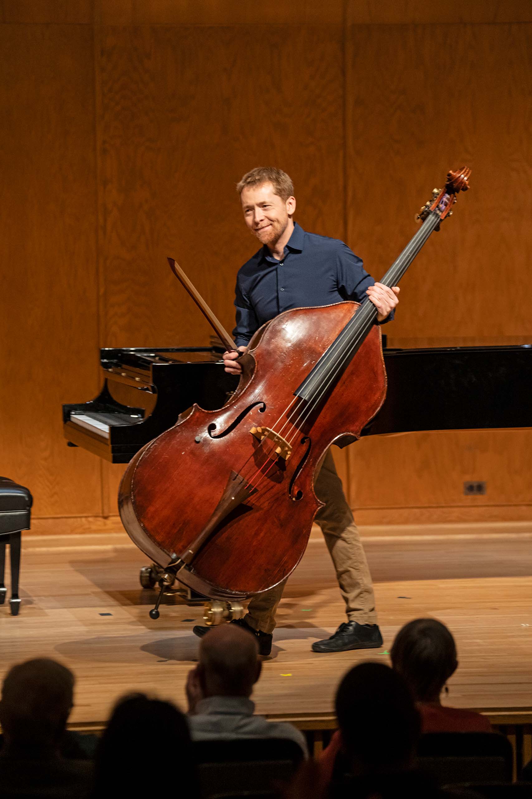 With audience heads visible in the lower foreground, Nicholas Walker stands on stage with a smile, holding his double bass and bow.