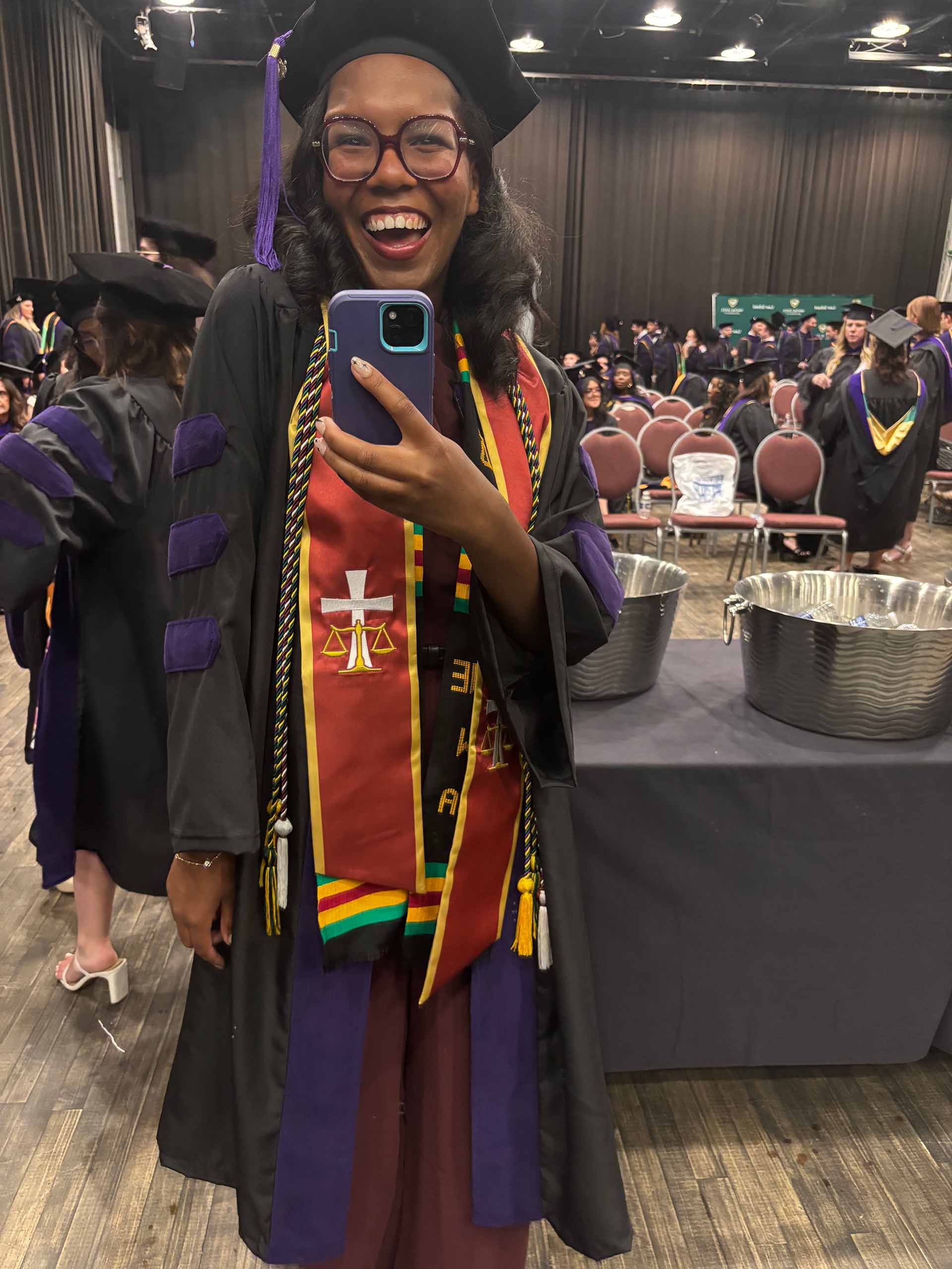 Wearing academic regalia with purple and red colors, Nya Johnson holds up her phone taking a selfie, with many other graduates seated and standing in the background.