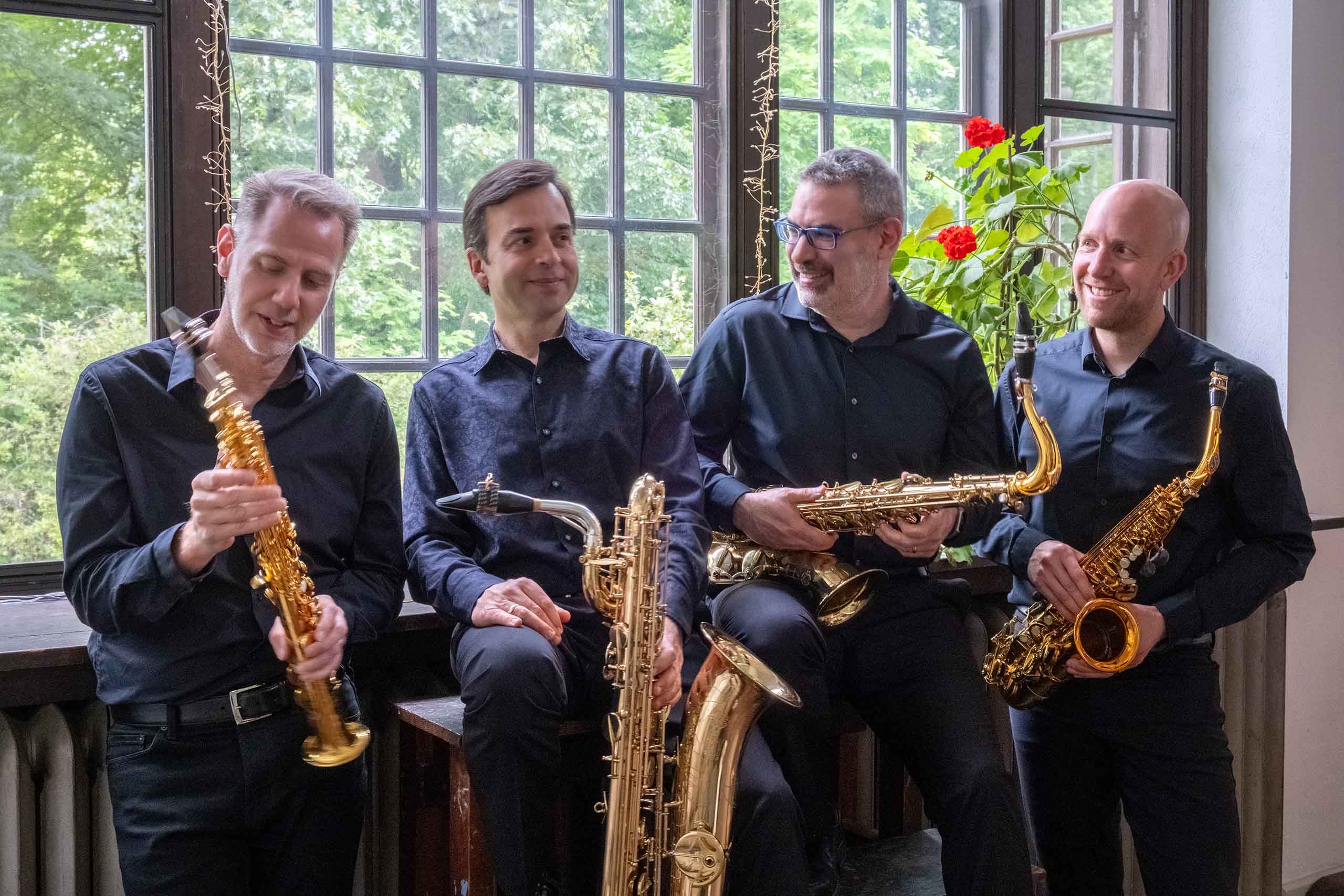 Candid photo of four people, wearing all black with collared shirts, and all holding gold saxophones. Paned windows looking over the woods, and a red geranium are behind them.