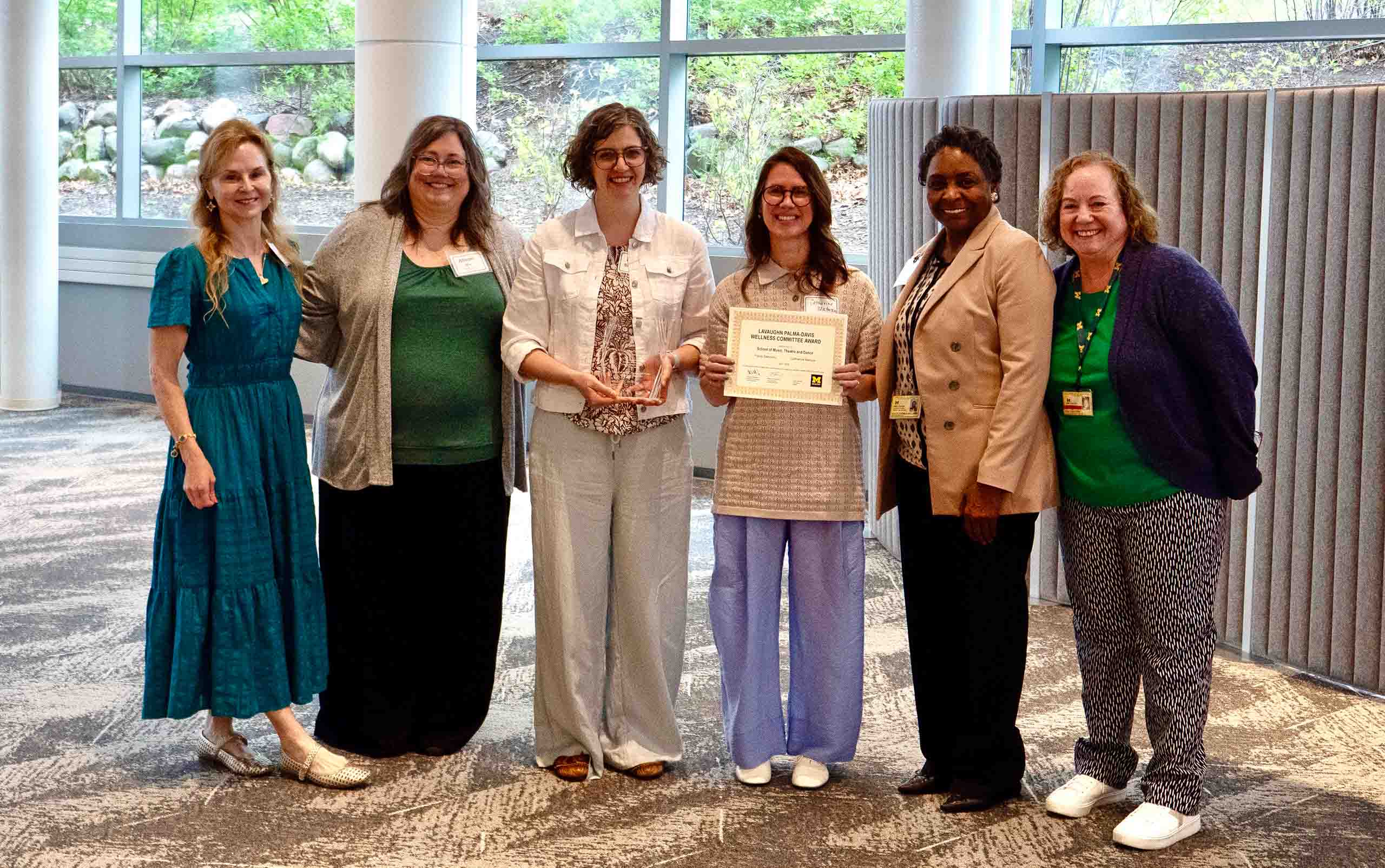 Six people pose standing together smiling, as one holds up a paper certificate and another holds a glass award. Behind them is a lobby space with a wall of windows facing on a bed of landscaping.