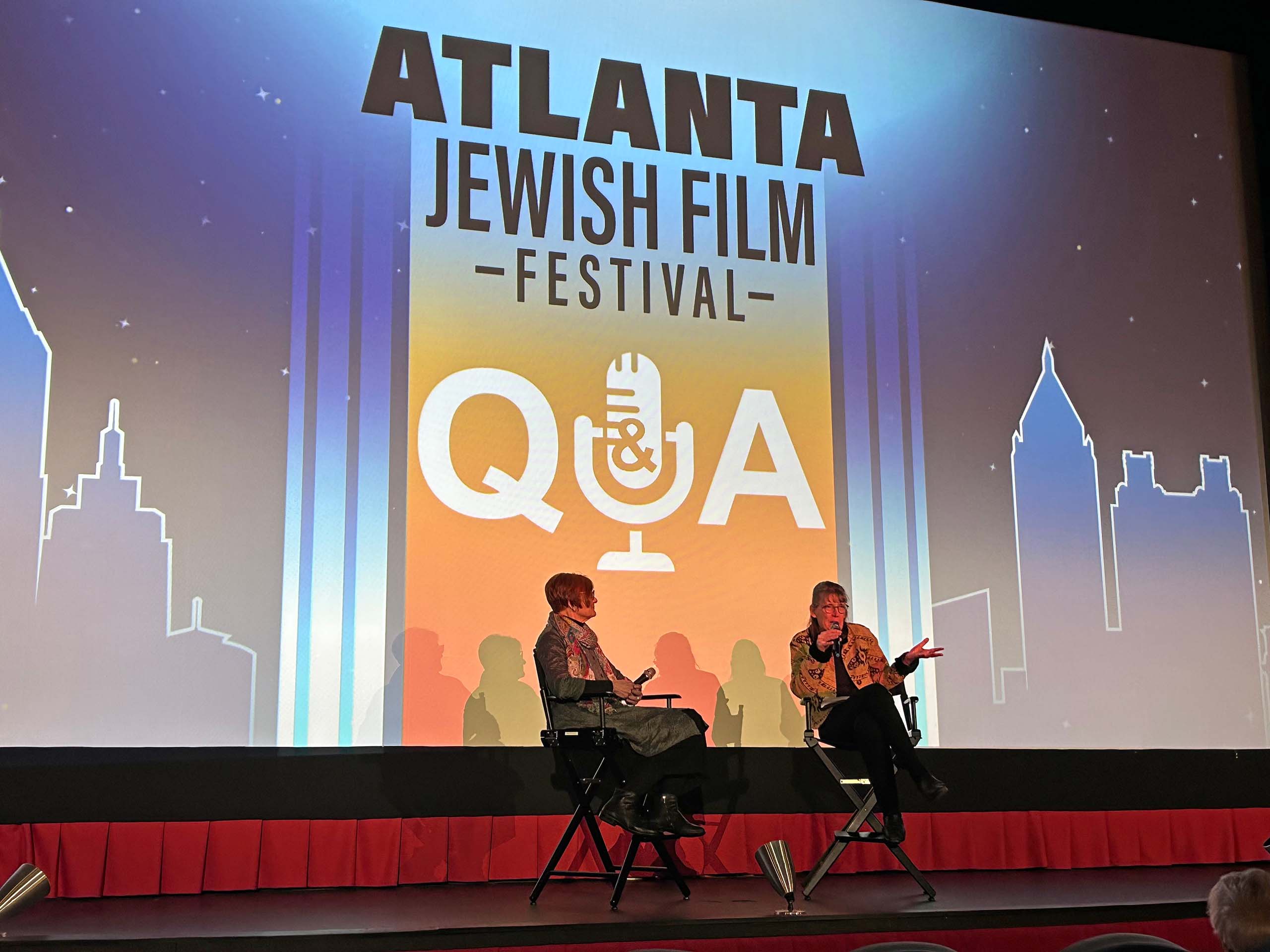 Two people seated in director chairs hold microphones on a stage. Behind them, a large projection says: Atlanta Jewish Film Festival Q&A with graphic of a city skyline and a microphone.