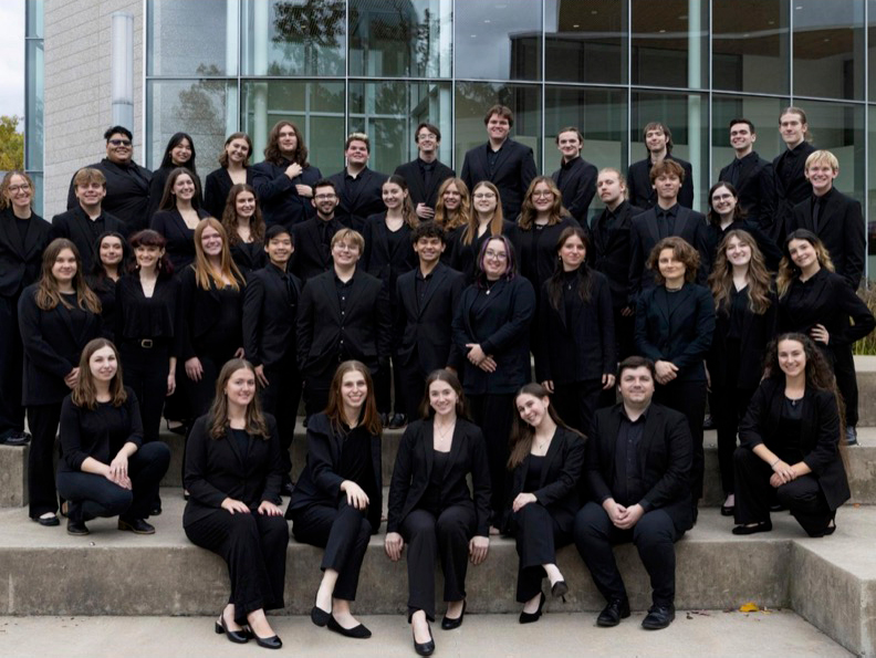 A mixed choir of over 3 dozen members poses in several rows outside a modern building with glass wall. All are attired in concert black. 