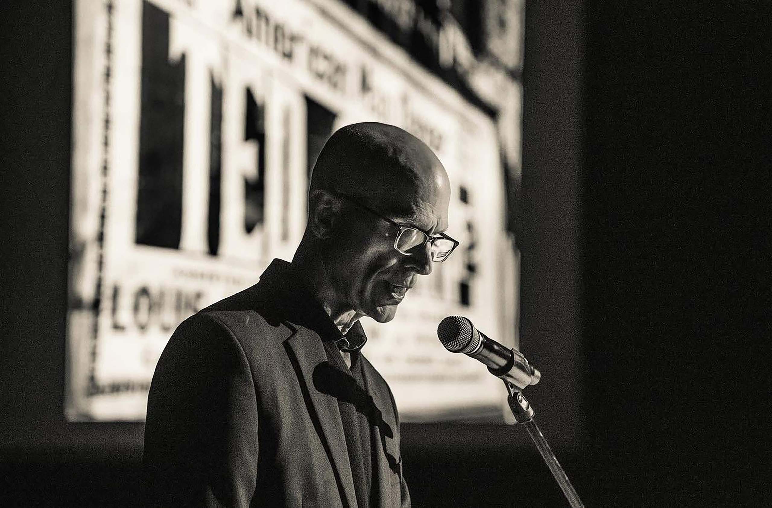 Silver-toned greyscale photo of Peter Sparling speaking into a microphone, with an obscured projection of a vintage-looking image behind him.