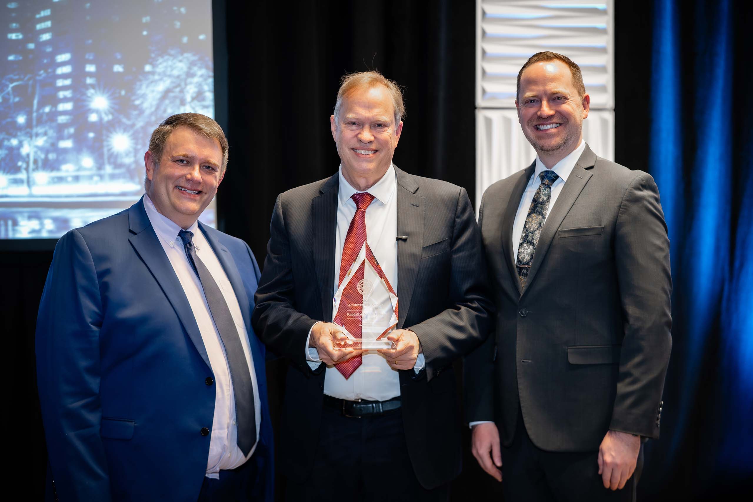 Three people pose standing together, all attired in suits and ties. Randall Faber is in the middle, holding up an engraved glass trophy.